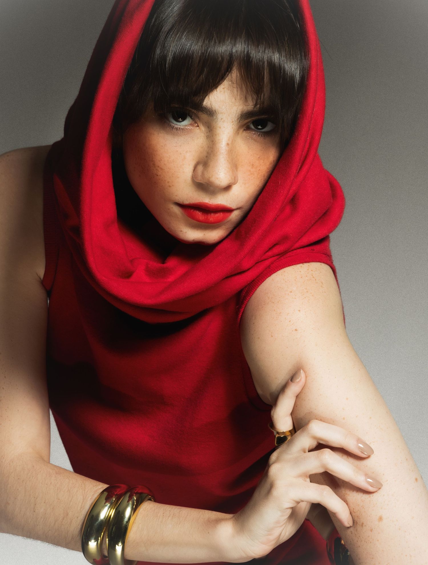 Fashion portrait of a freckled woman with dark bangs in a red hooded cowl top, red lipstick and gold bangles.