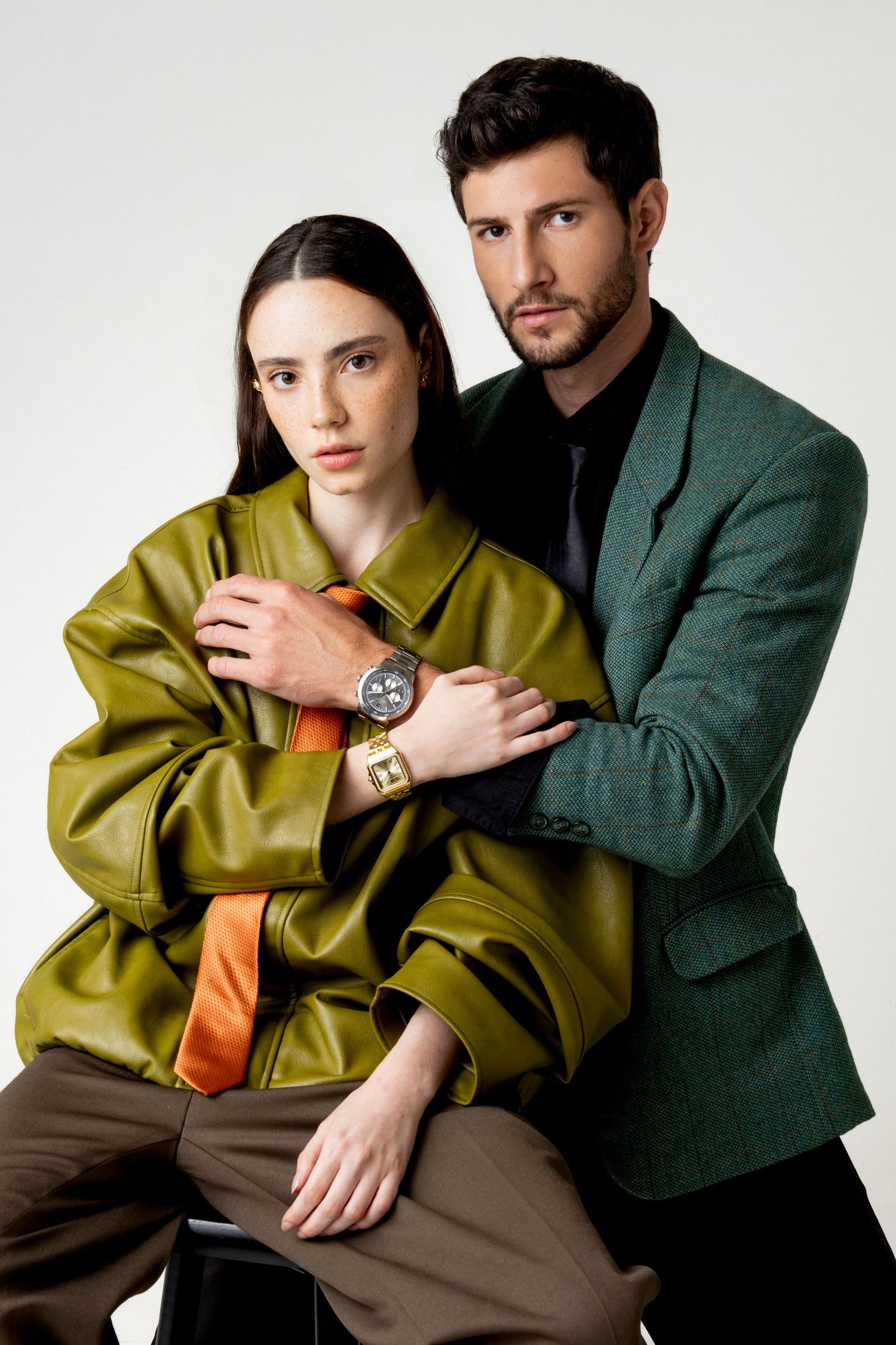 Woman in an olive leather jacket and orange tie sits as a bearded man in a green blazer embraces her, both wearing wristwatches.