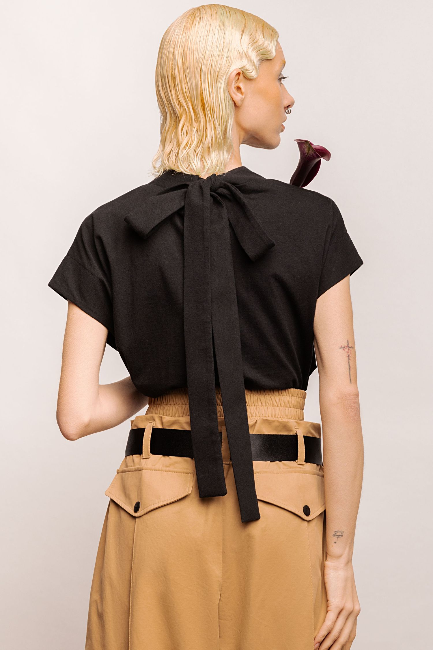 Back view of a woman in a black short-sleeve top with a long tie bow at the neck and a high-waisted tan skirt with a black belt.