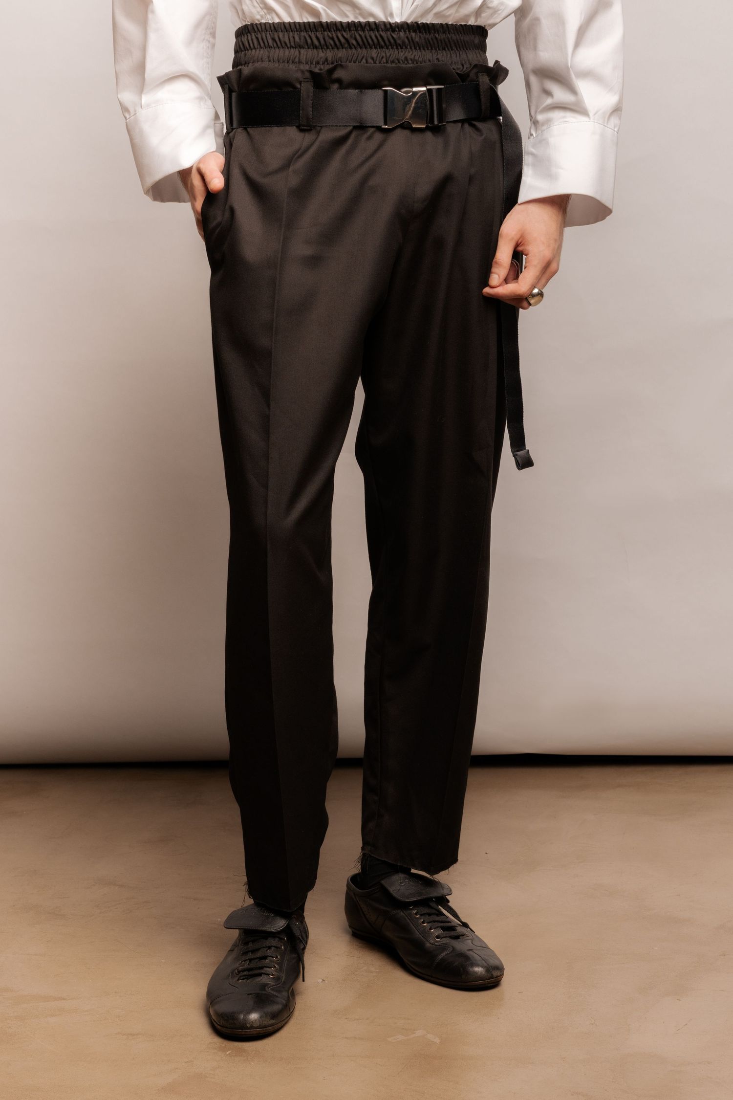 Close-up of black high-waisted trousers with an elasticated waistband, buckled strap belt and black leather lace-up shoes.
