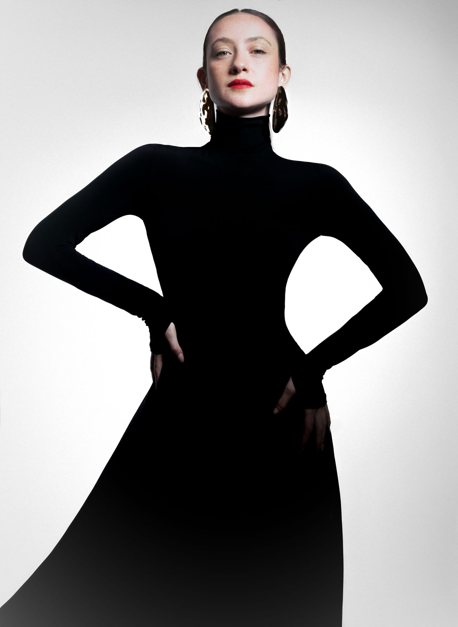 Woman in a black turtleneck dress with hands on hips and gold earrings poses against a white studio background.