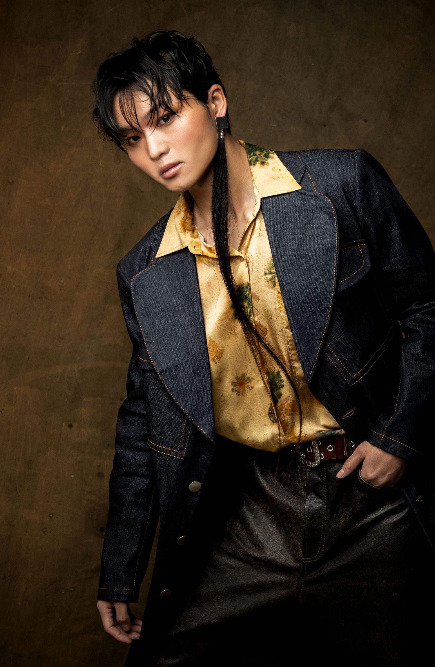 Close-up of a model in a denim jacket and gold floral shirt with a long braid, looking at the camera against a brown backdrop.