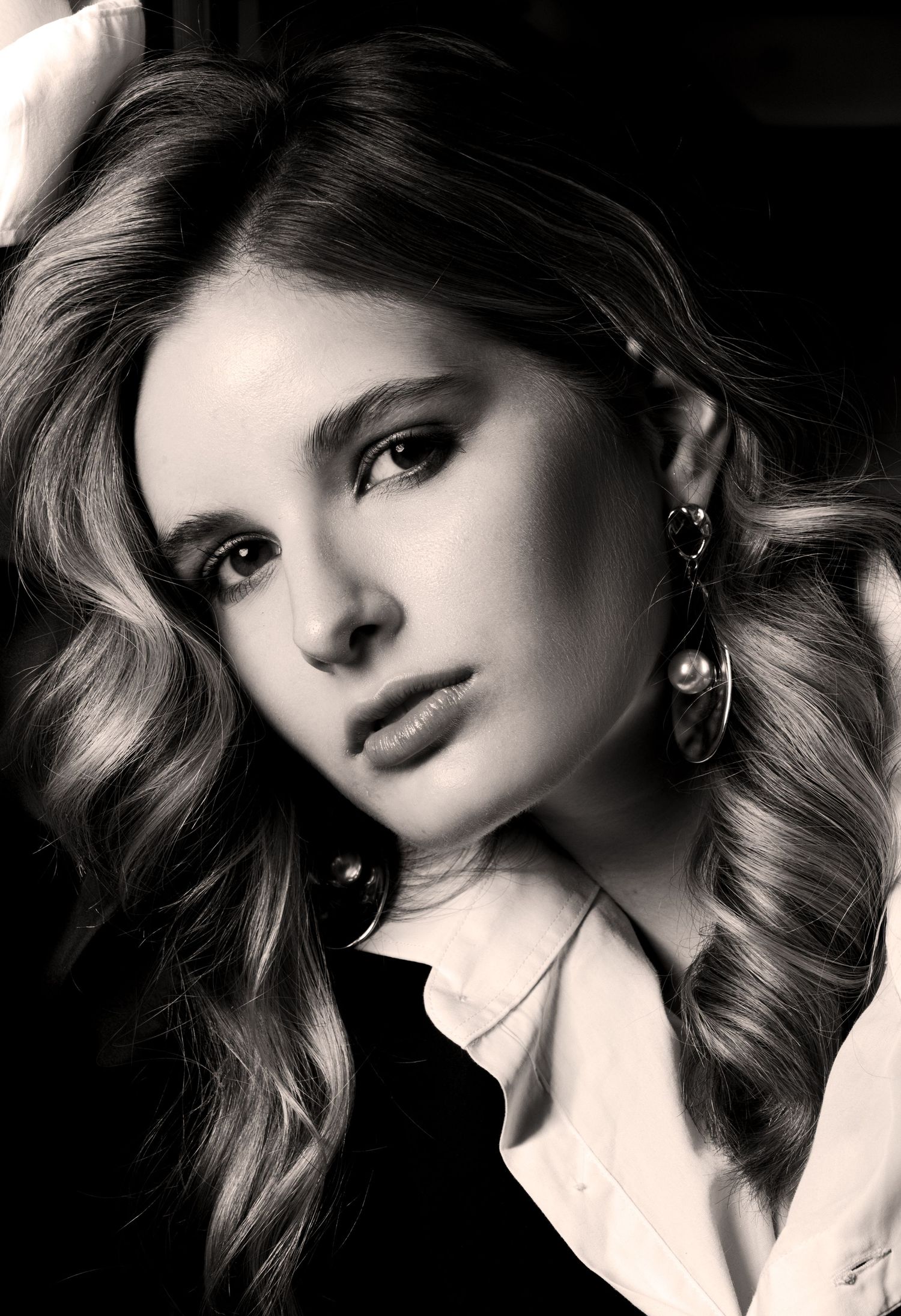 Black and white close-up portrait of a model with long wavy hair, pearl drop earrings and a white collared shirt.