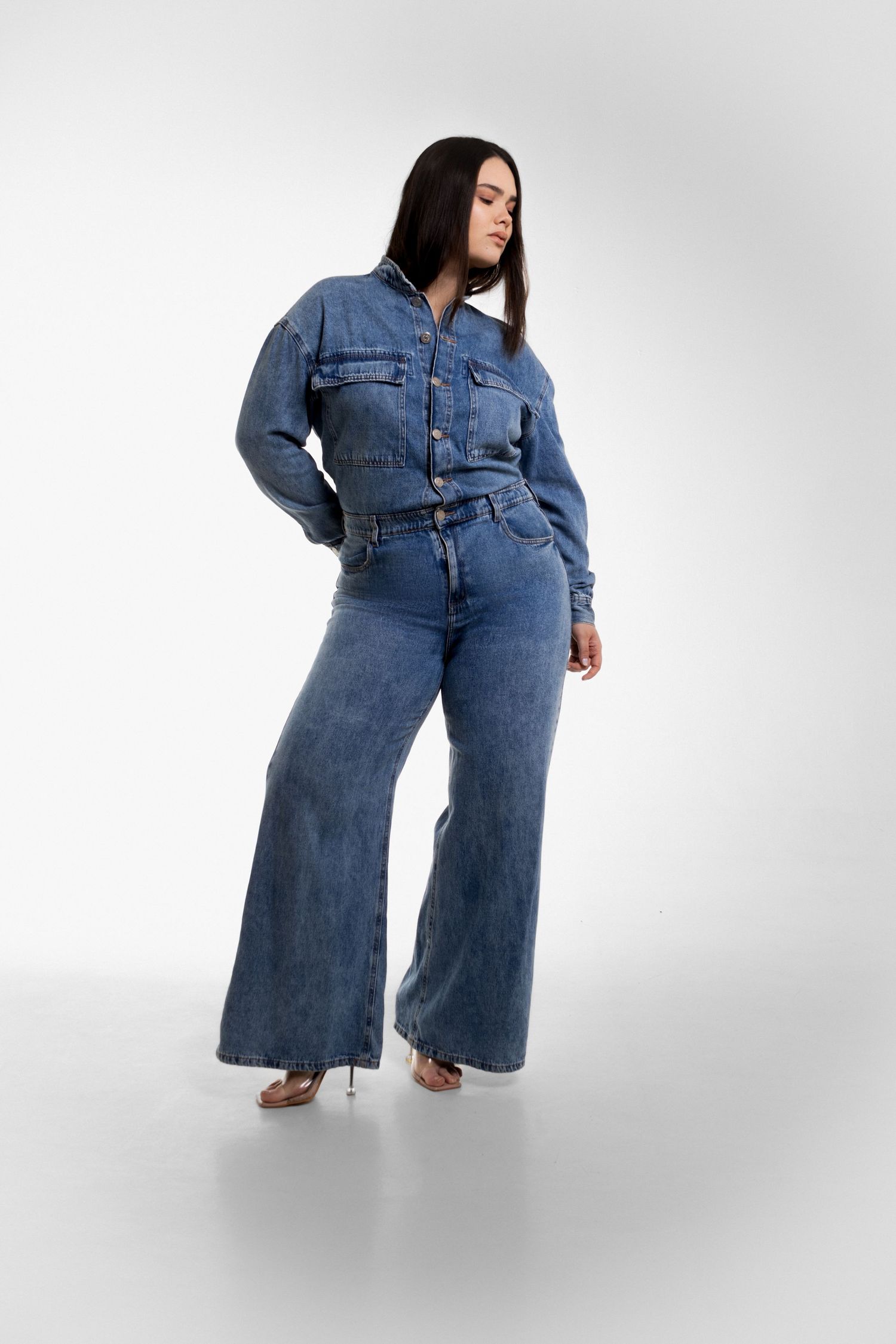 Full-length studio shot of a model in a blue denim jumpsuit with wide-leg pants and clear heeled sandals against a white background.
