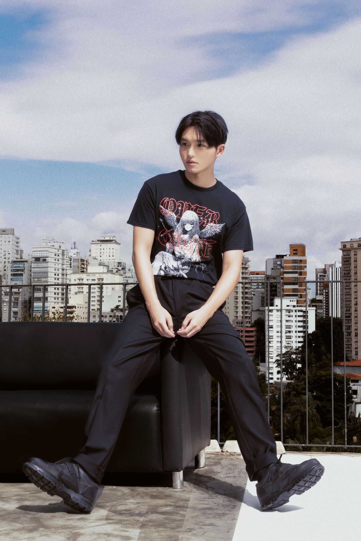 A young man in a black anime graphic t-shirt and black trousers sits on a rooftop sofa overlooking a sunlit city skyline.