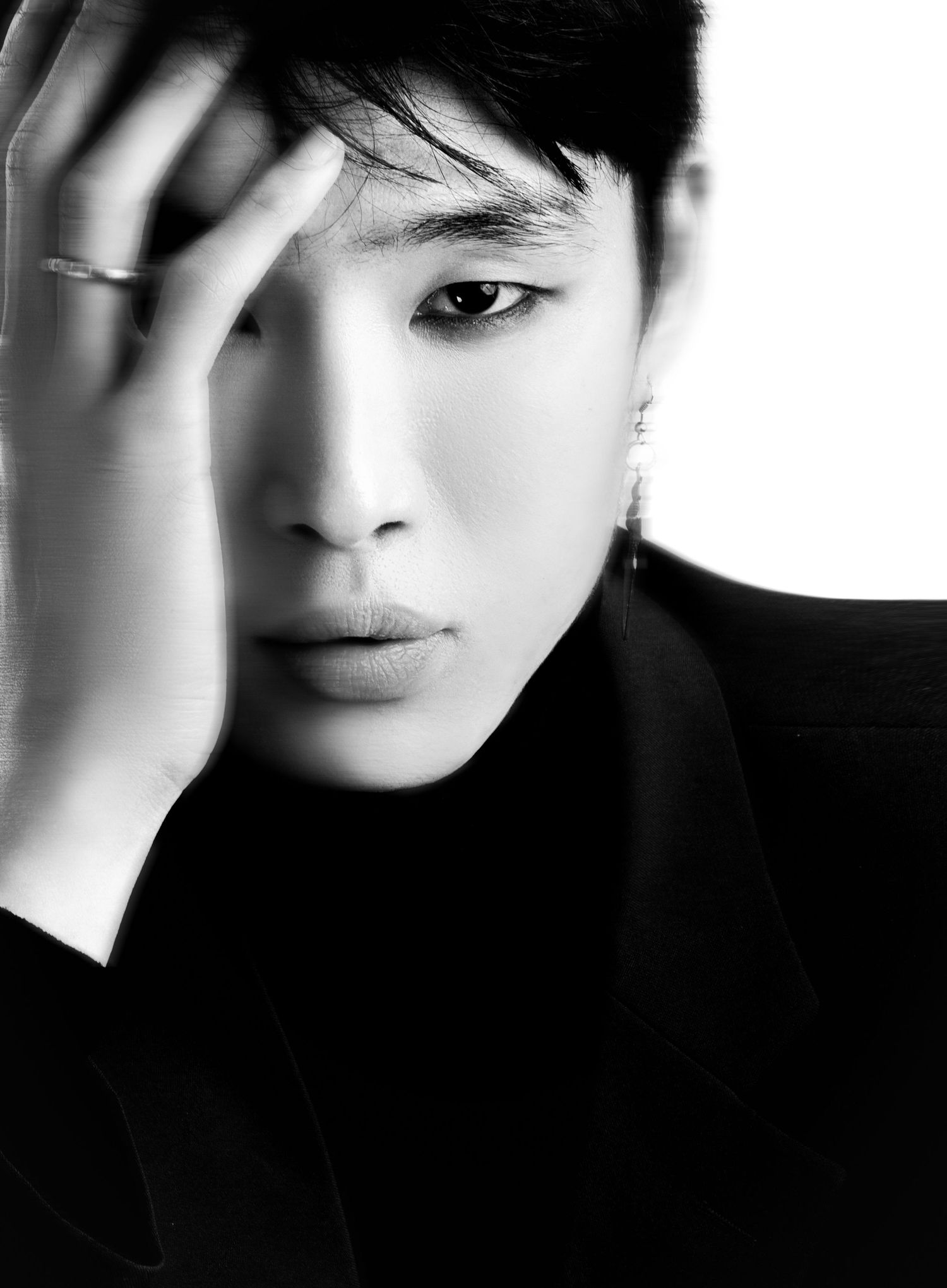 Black-and-white portrait of a model with dark hair, hand raised to the forehead, wearing a ring, an earring and a dark high-neck top.
