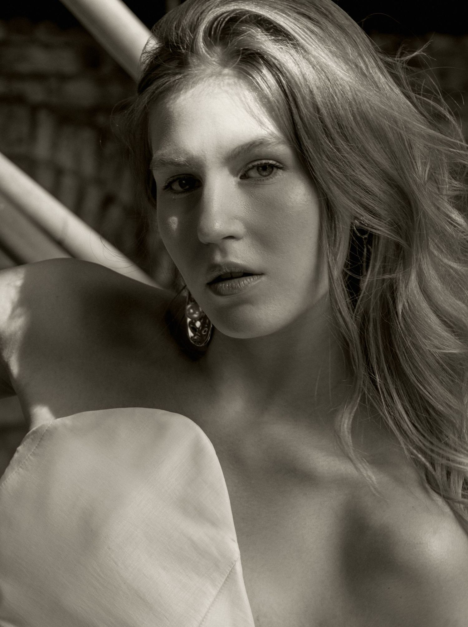 Black and white close-up portrait of a model with long wavy hair, a statement earring and a bare shoulder draped in soft light.