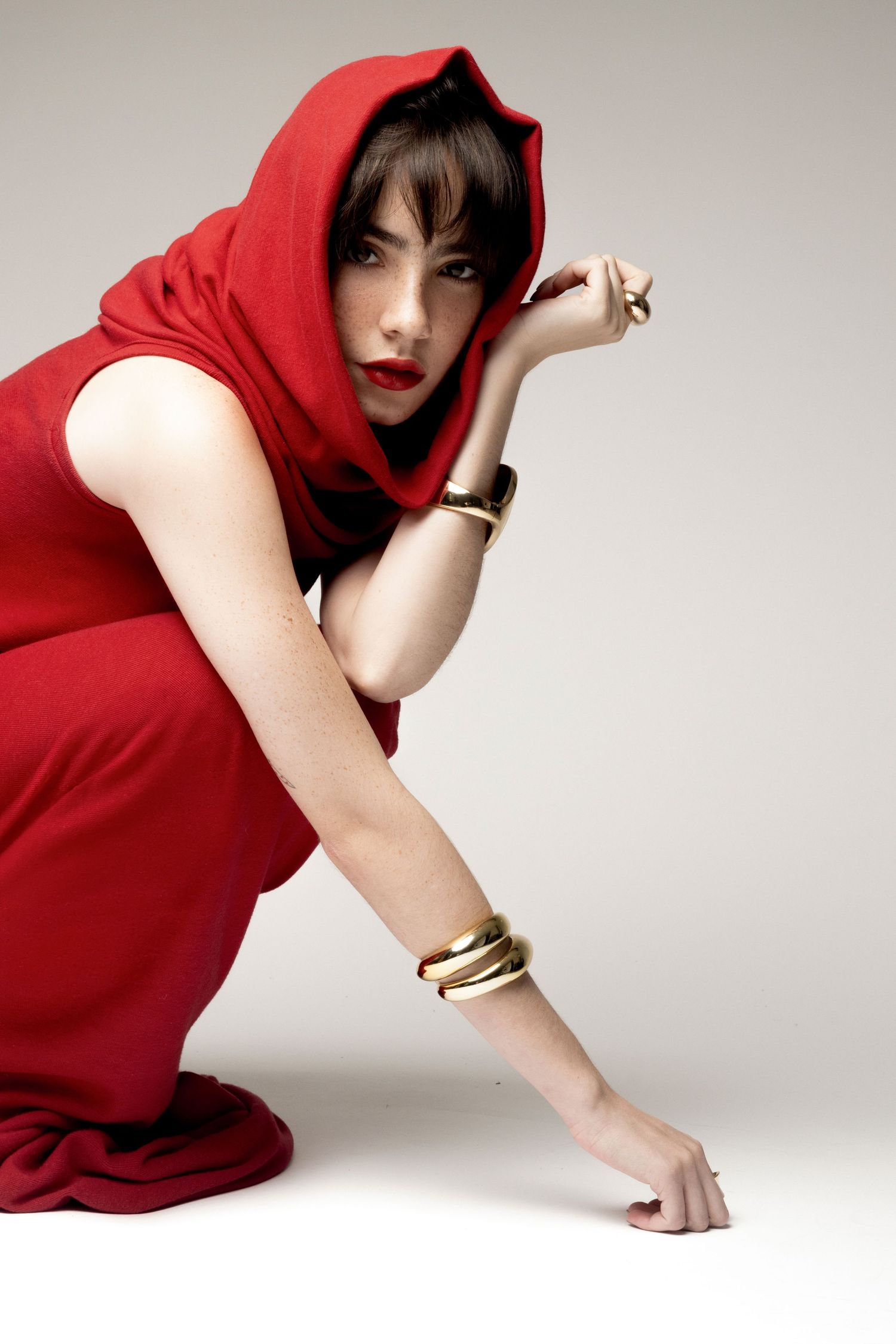 Woman in a red hooded dress crouches in a studio, wearing stacked gold bangles and rings with a dramatic gaze.