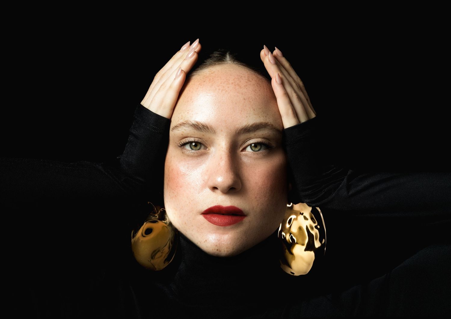 Close portrait of a woman with red lips and oversized gold earrings, hands raised beside her head on a black backdrop.