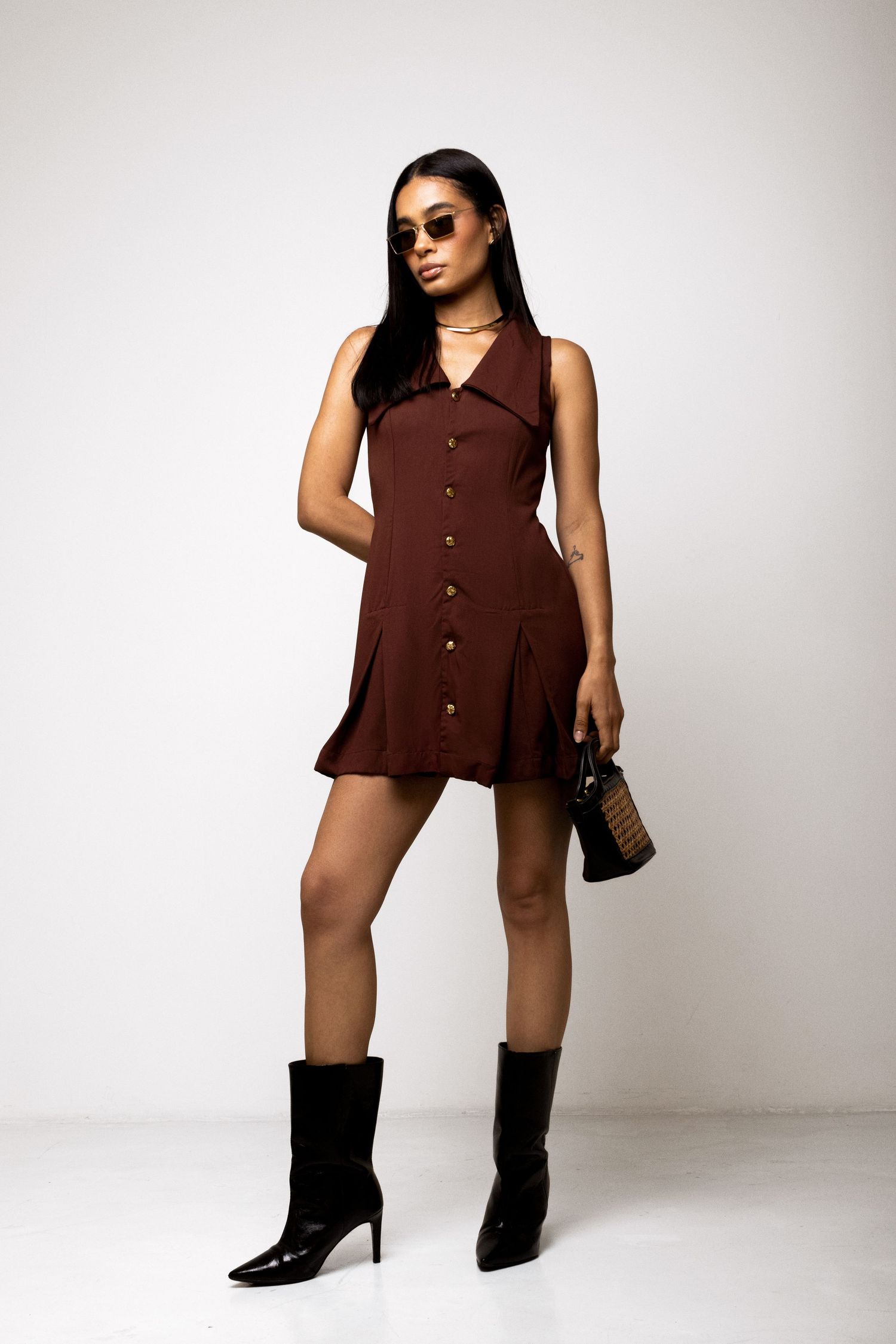 Woman in a brown sleeveless button-front mini dress with wide collar, black pointed ankle boots, sunglasses and a woven handbag.