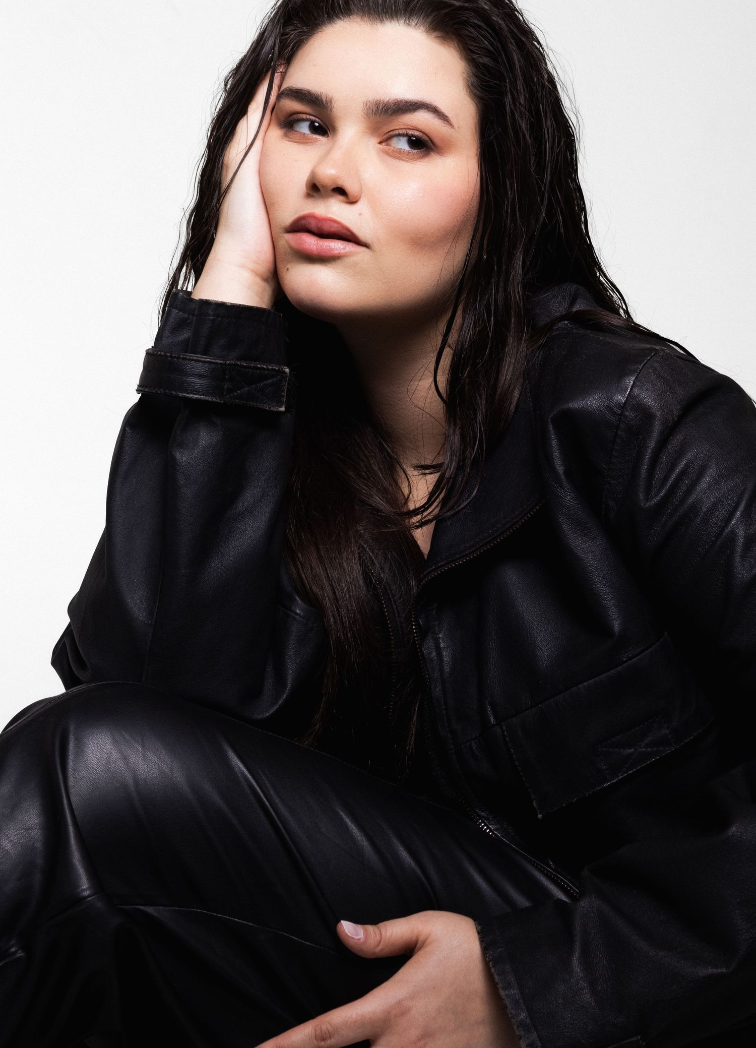 Model with wet-look dark hair rests her cheek on her hand, dressed in a black leather jacket and leather pants.