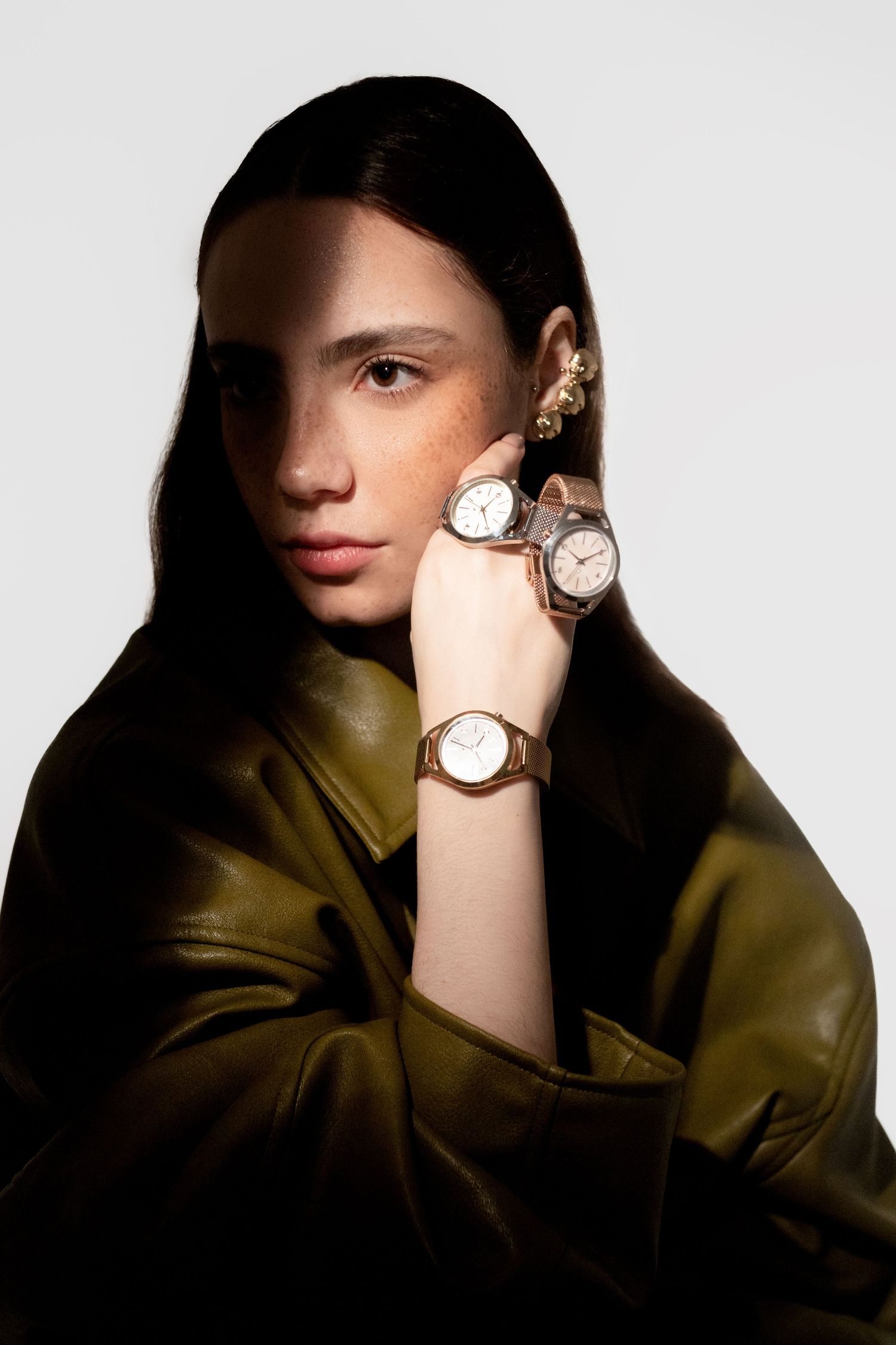 Woman in an olive leather jacket poses with three wristwatches on her raised hand and wrist against a plain backdrop.