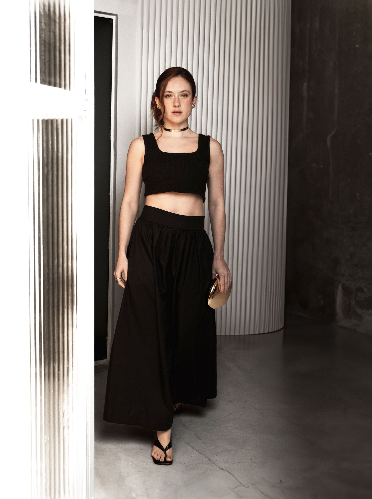 Woman in a black crop top and flowing black maxi skirt with sandals stands in a modern space with ribbed white walls.