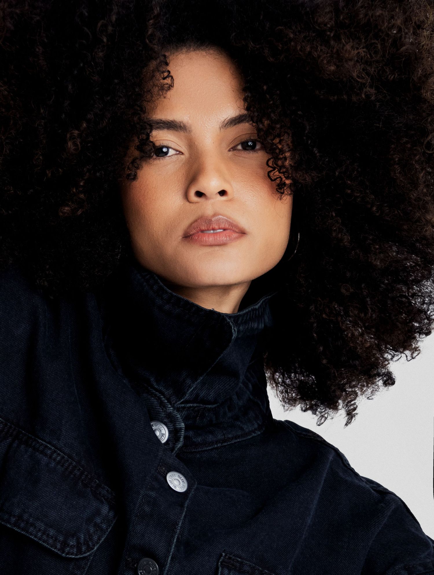 Close-up portrait of a model with full, voluminous curly hair wearing a black denim jacket with silver buttons and a confident gaze.