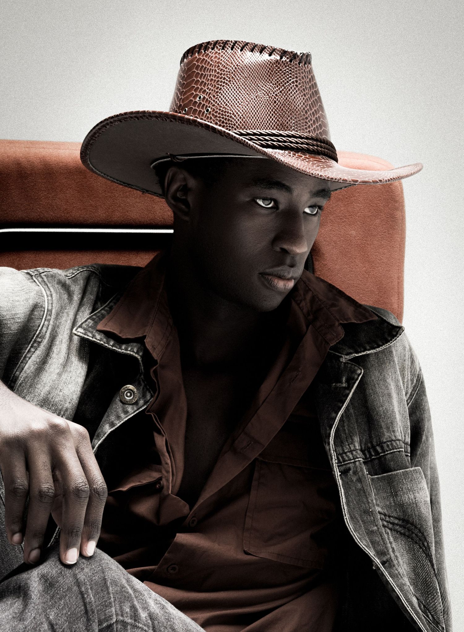 Male model in a brown snakeskin-textured cowboy hat, rust shirt and gray denim jacket sits in a rust-colored chair with a sideways glance.
