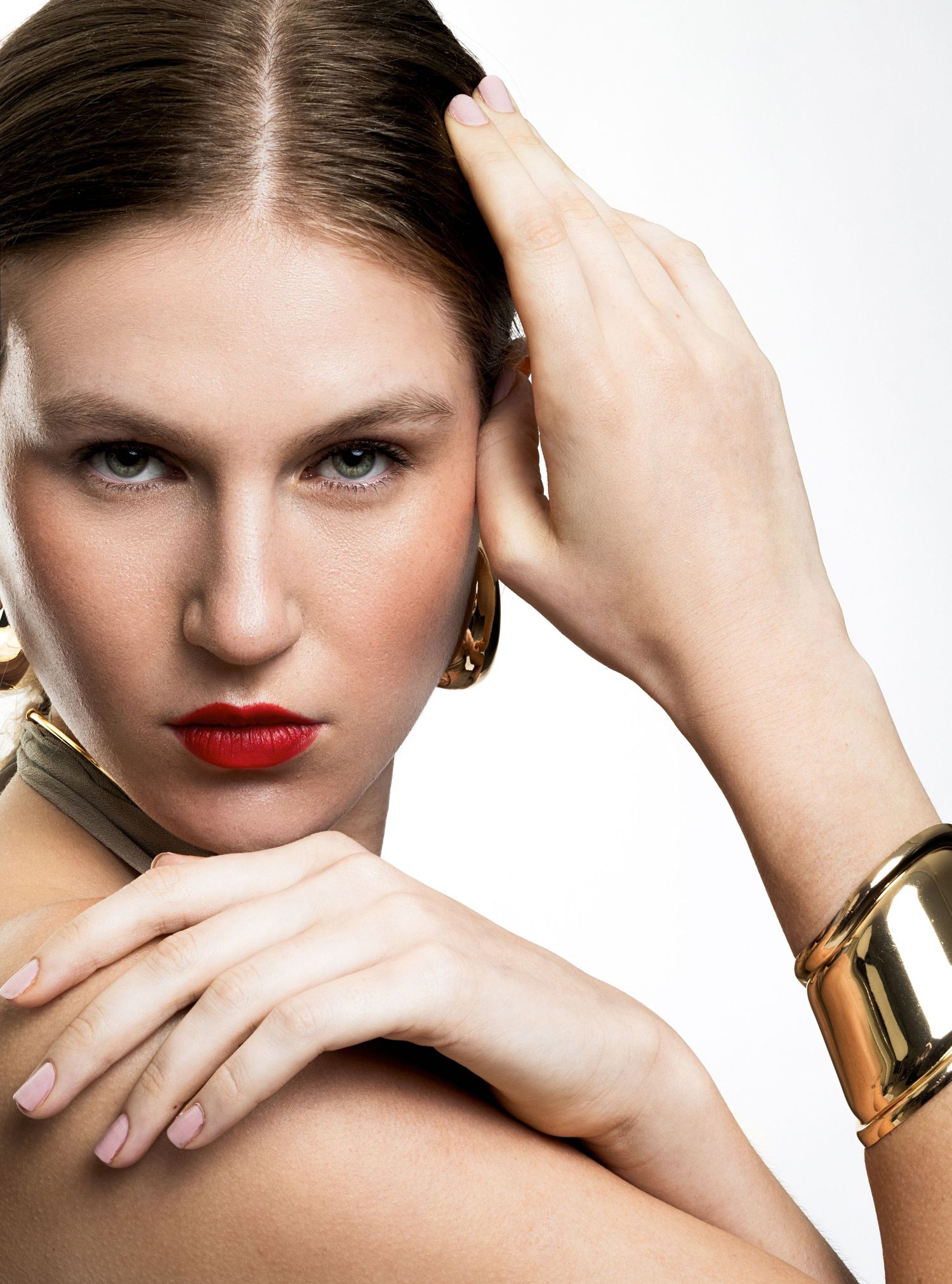 Close-up beauty portrait of a model with red lipstick, a gold cuff bracelet and gold hoop earrings, hands framing her face.
