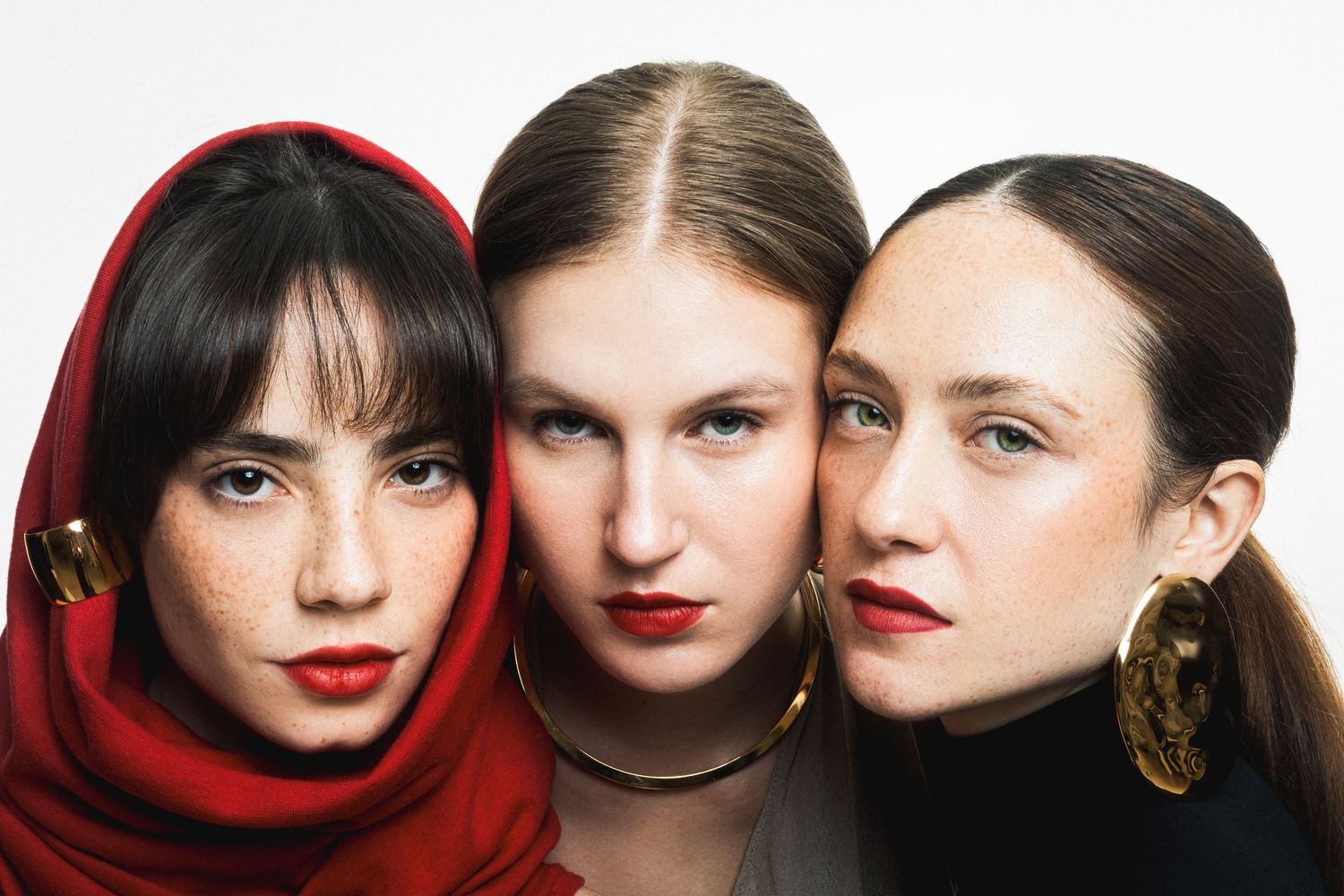 Three women with bold red lips pose side by side, one in a red headscarf and one with large gold earrings, against white.