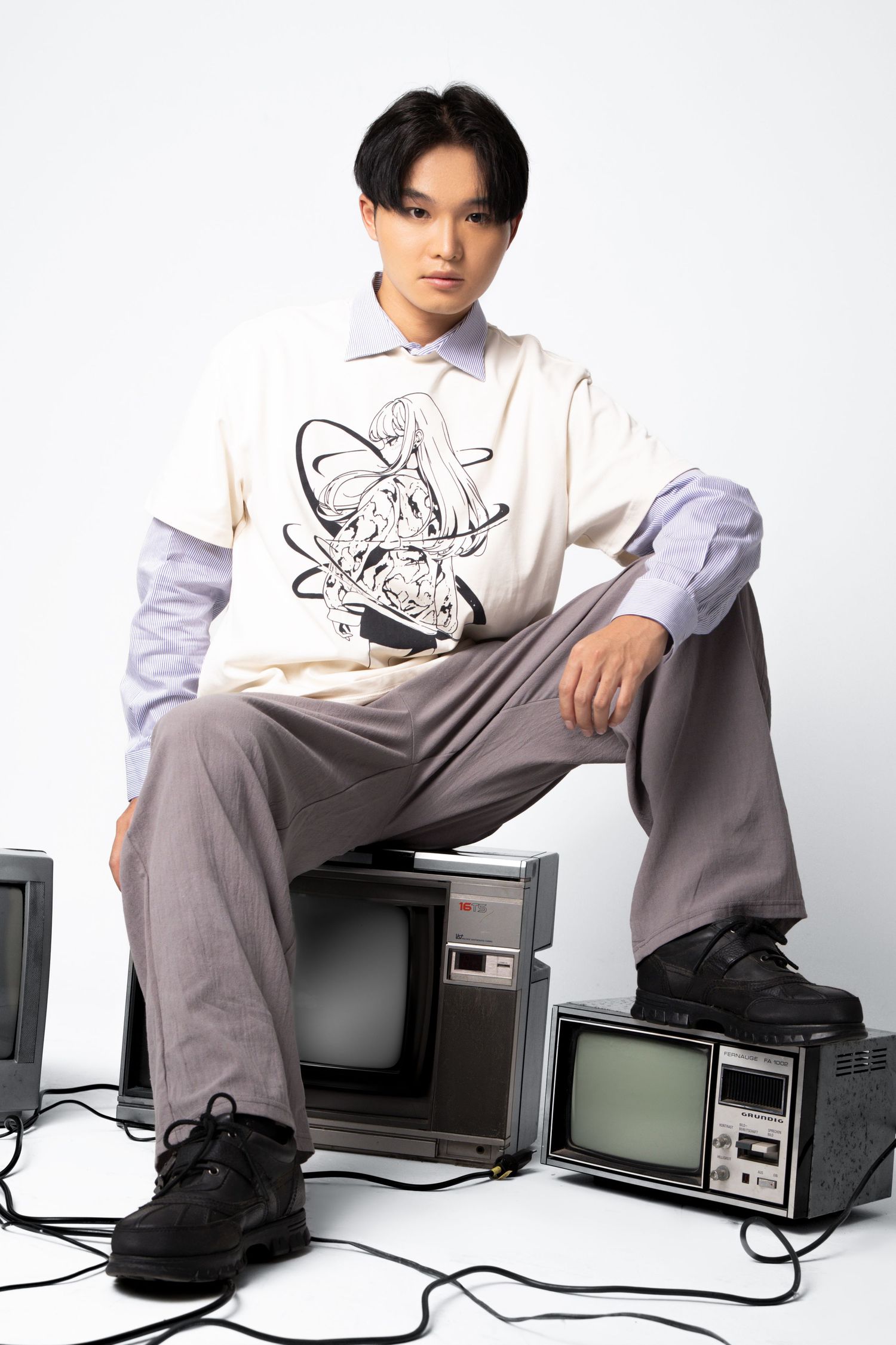 A model in a graphic cream tee and grey trousers sits on retro televisions with tangled cables on a white studio background.