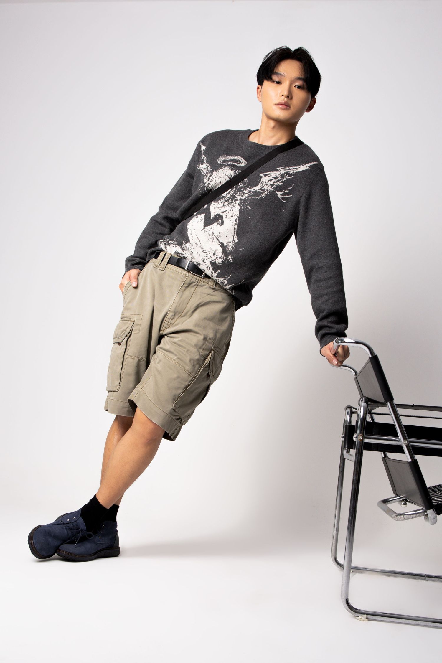 Model in a graphic sweater and khaki cargo shorts leans against a modern steel chair with a hand in his pocket.