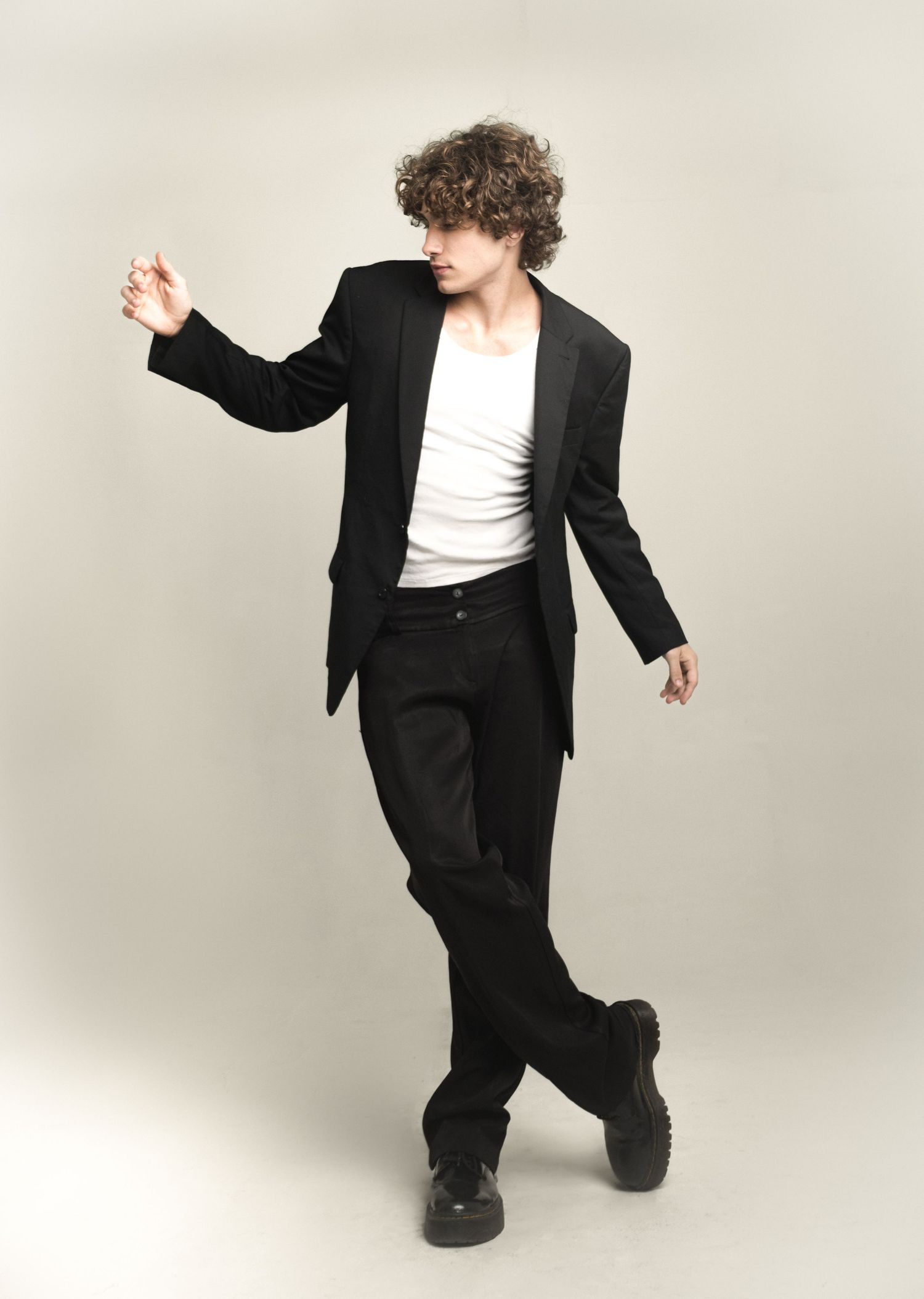 Model with curly hair poses in a black blazer, white tank top and wide black trousers with chunky shoes against a neutral backdrop.