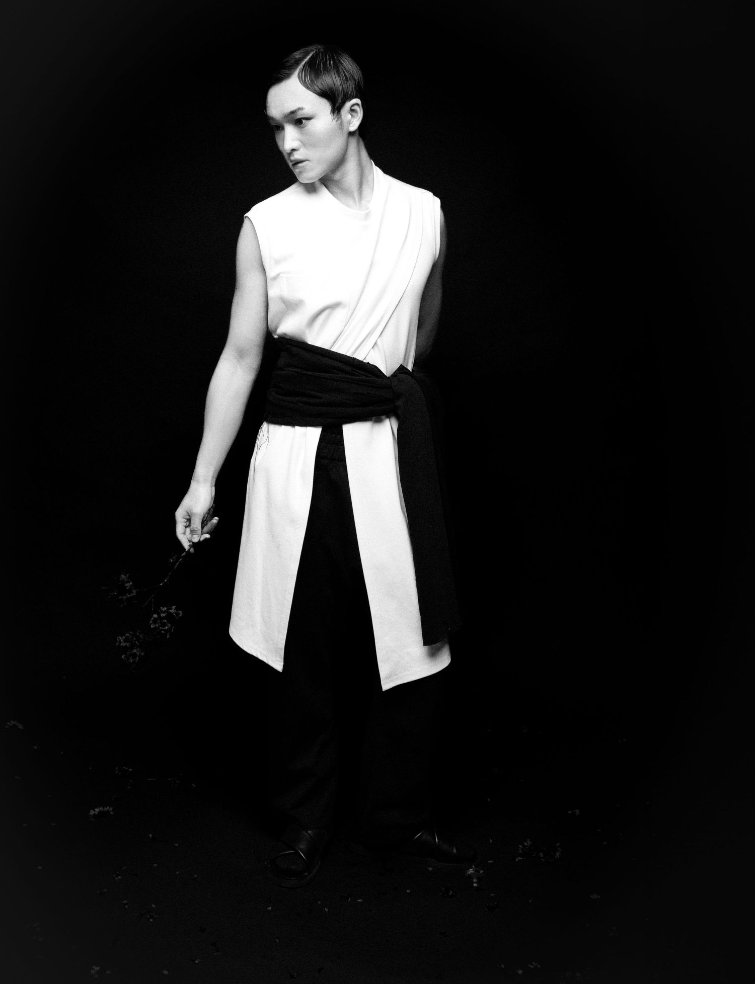 Black and white full-length portrait of a model in a sleeveless white tunic with a black sash, standing against a dark background.