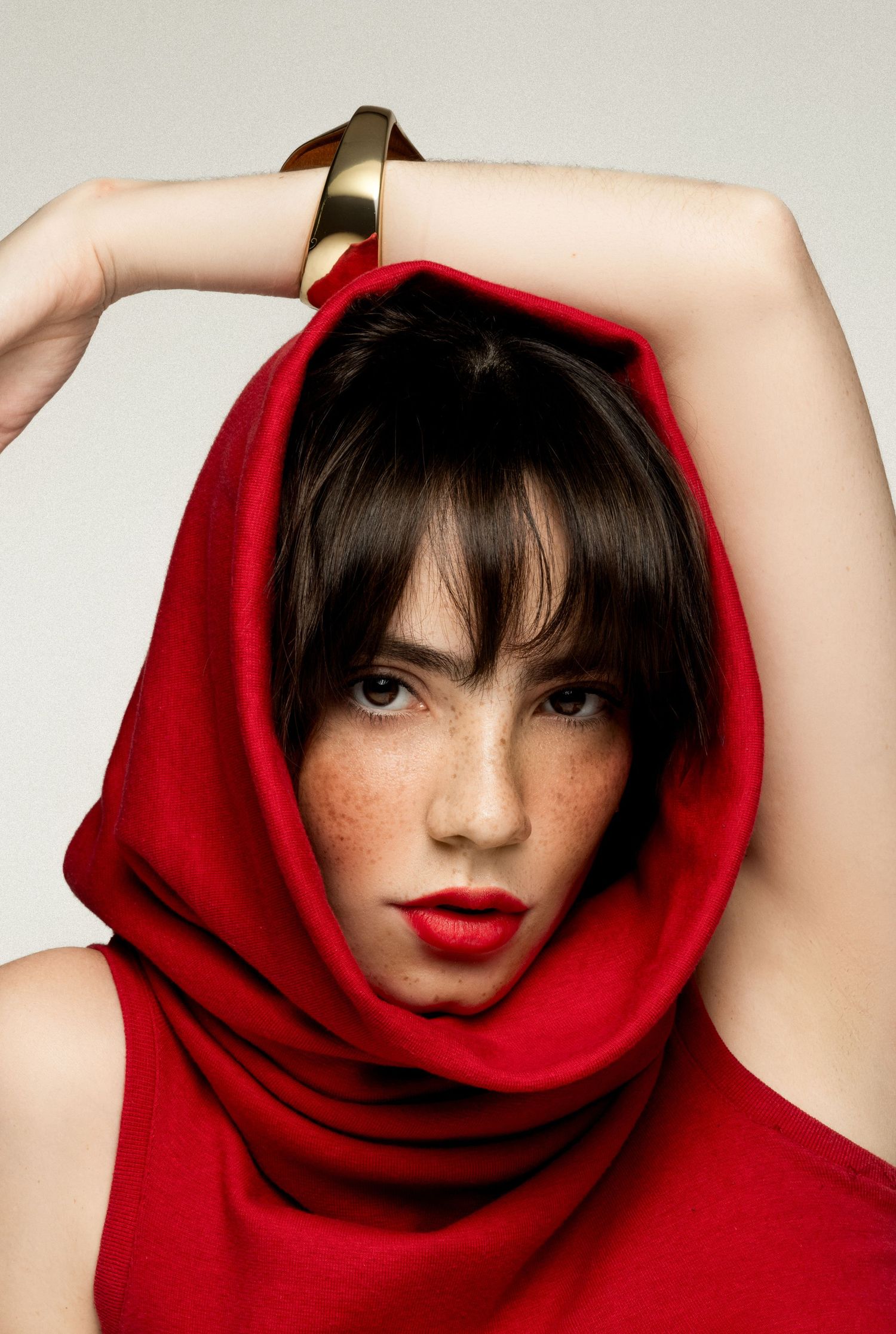 Close-up beauty portrait of a freckled woman in a red hood cowl, red lips, one arm raised with a gold bangle.