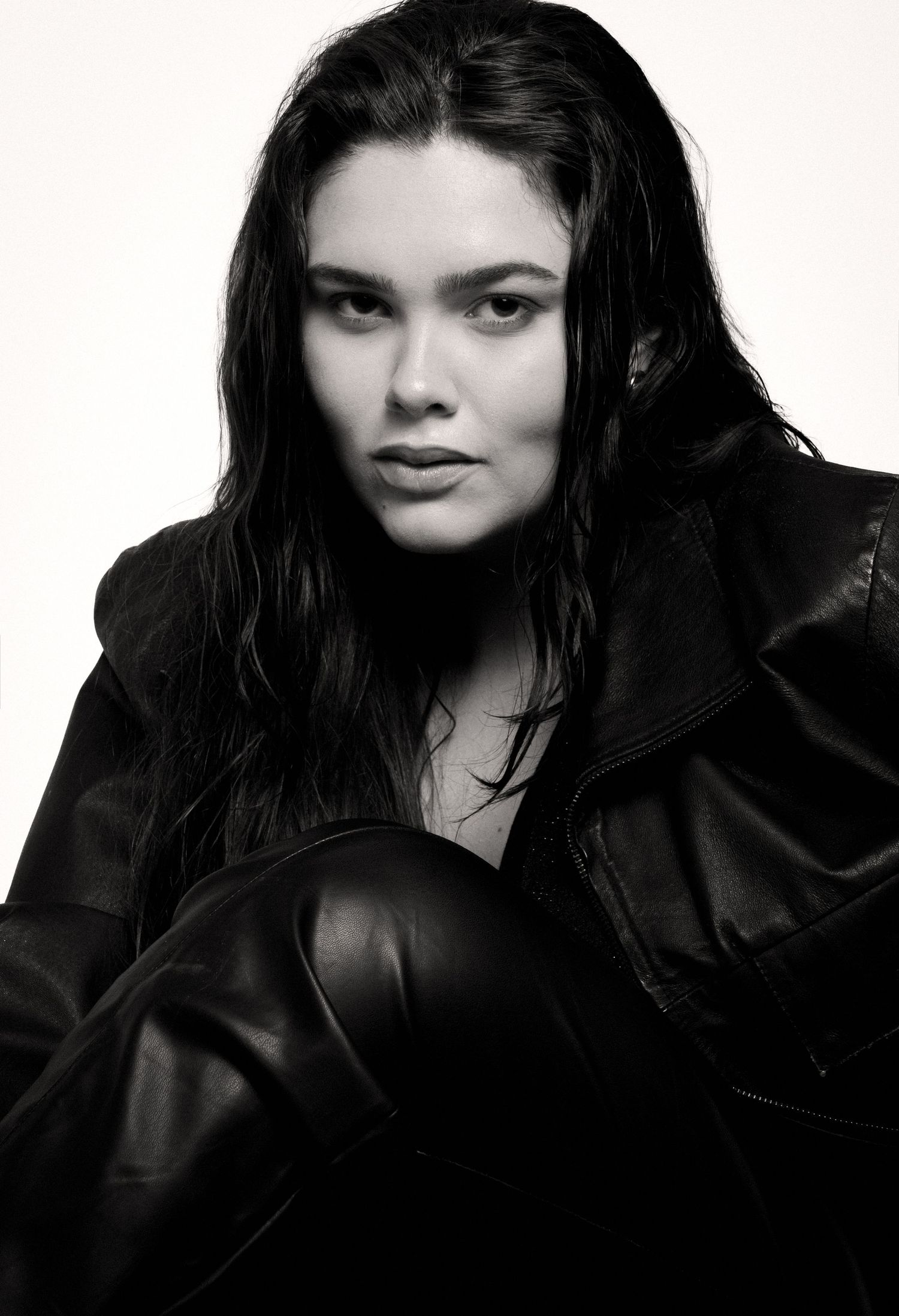 Black and white close-up of a model with wet-look long hair in a black leather jacket and leather pants, seated with a moody expression.