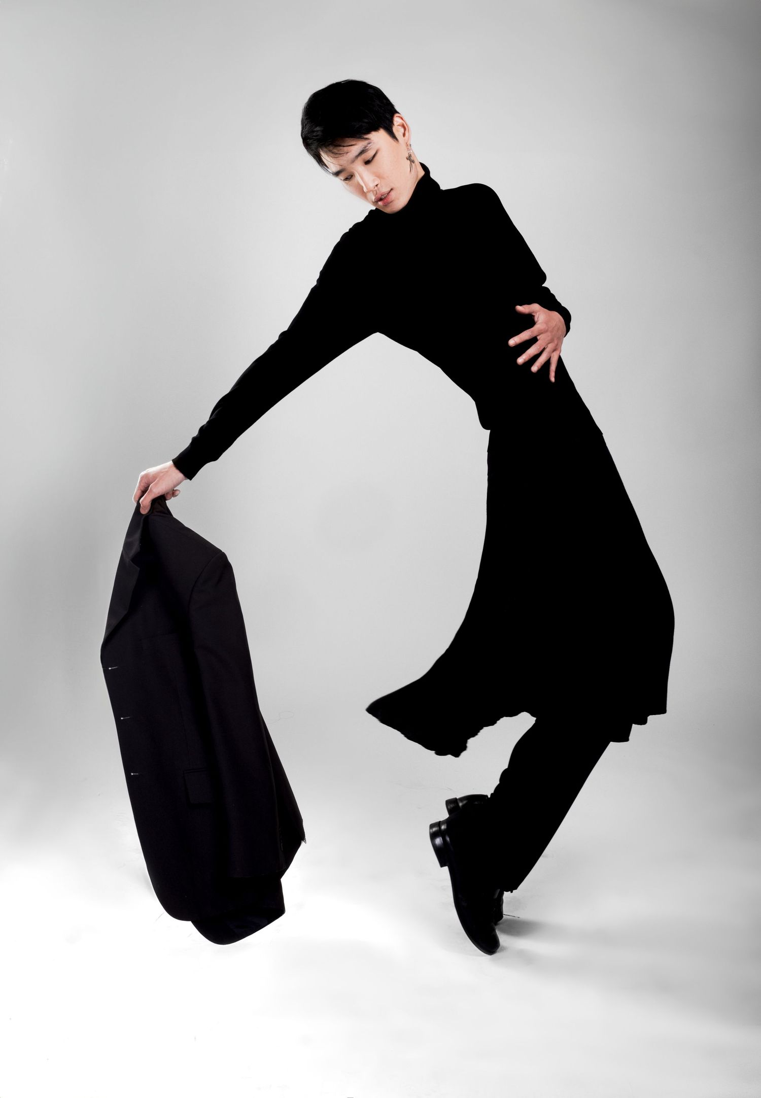 Model with short dark hair poses mid-movement in an all-black outfit, holding a black blazer in one hand against a gray backdrop.