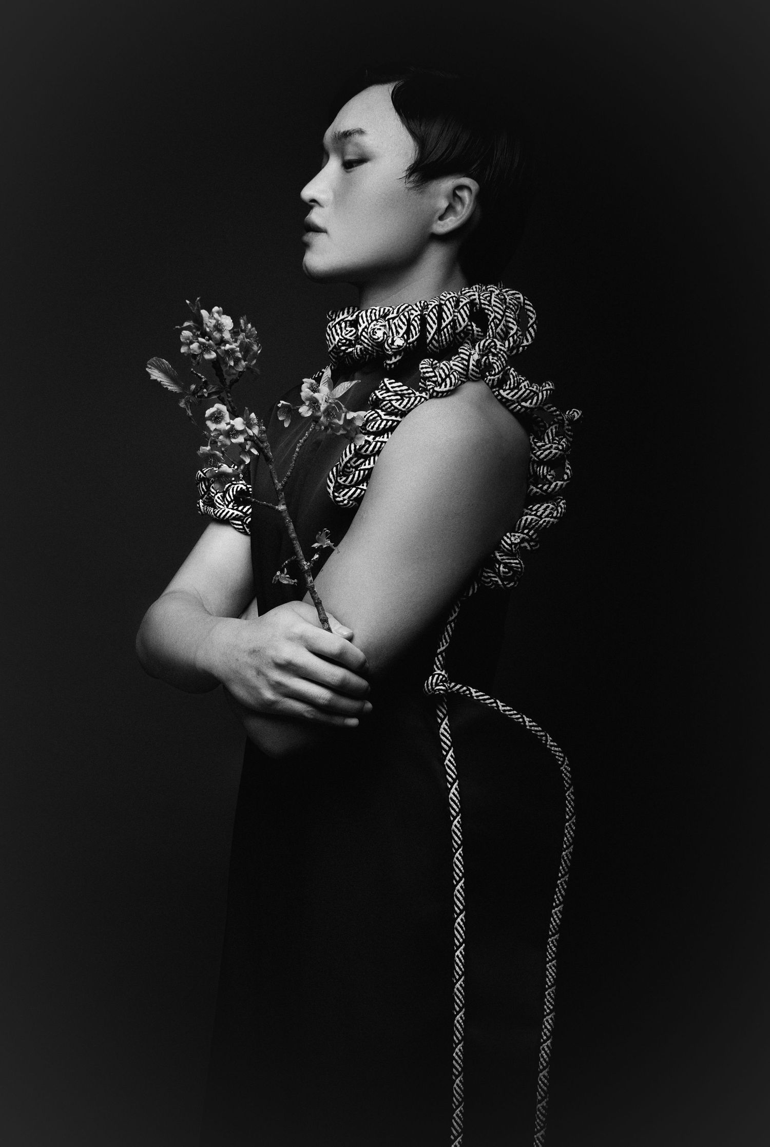 Black and white profile portrait of a model in a rope-textured ruffled collar dress holding a flowering branch against a dark background.