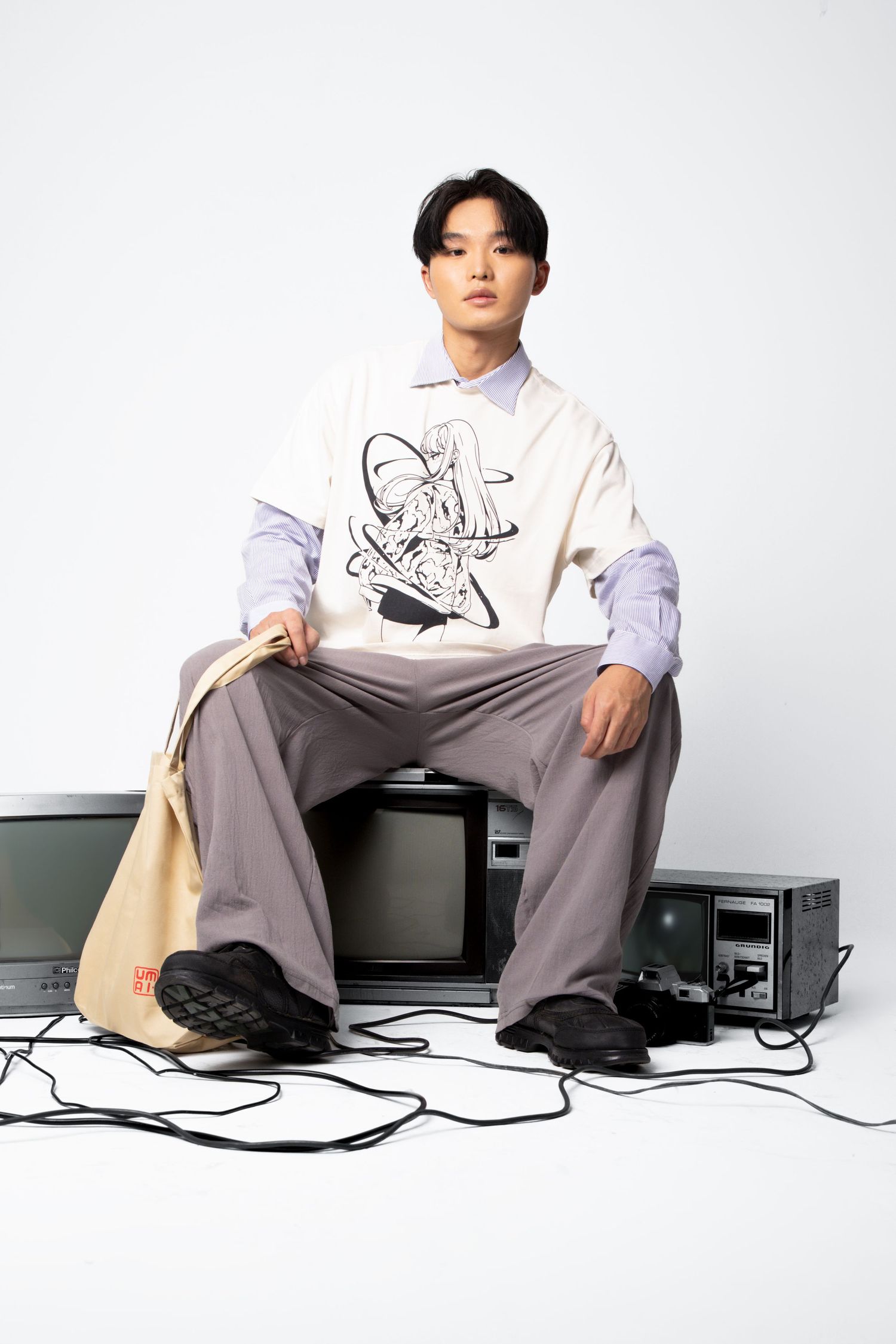 A model in a cream graphic vest and grey trousers sits on an old television holding a beige tote bag in the studio.