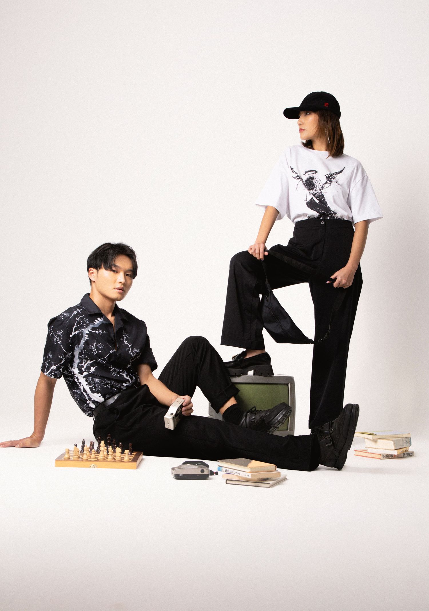 A man in a black patterned shirt sits by a chess set and books while a woman in a cap and white graphic tee stands behind him.