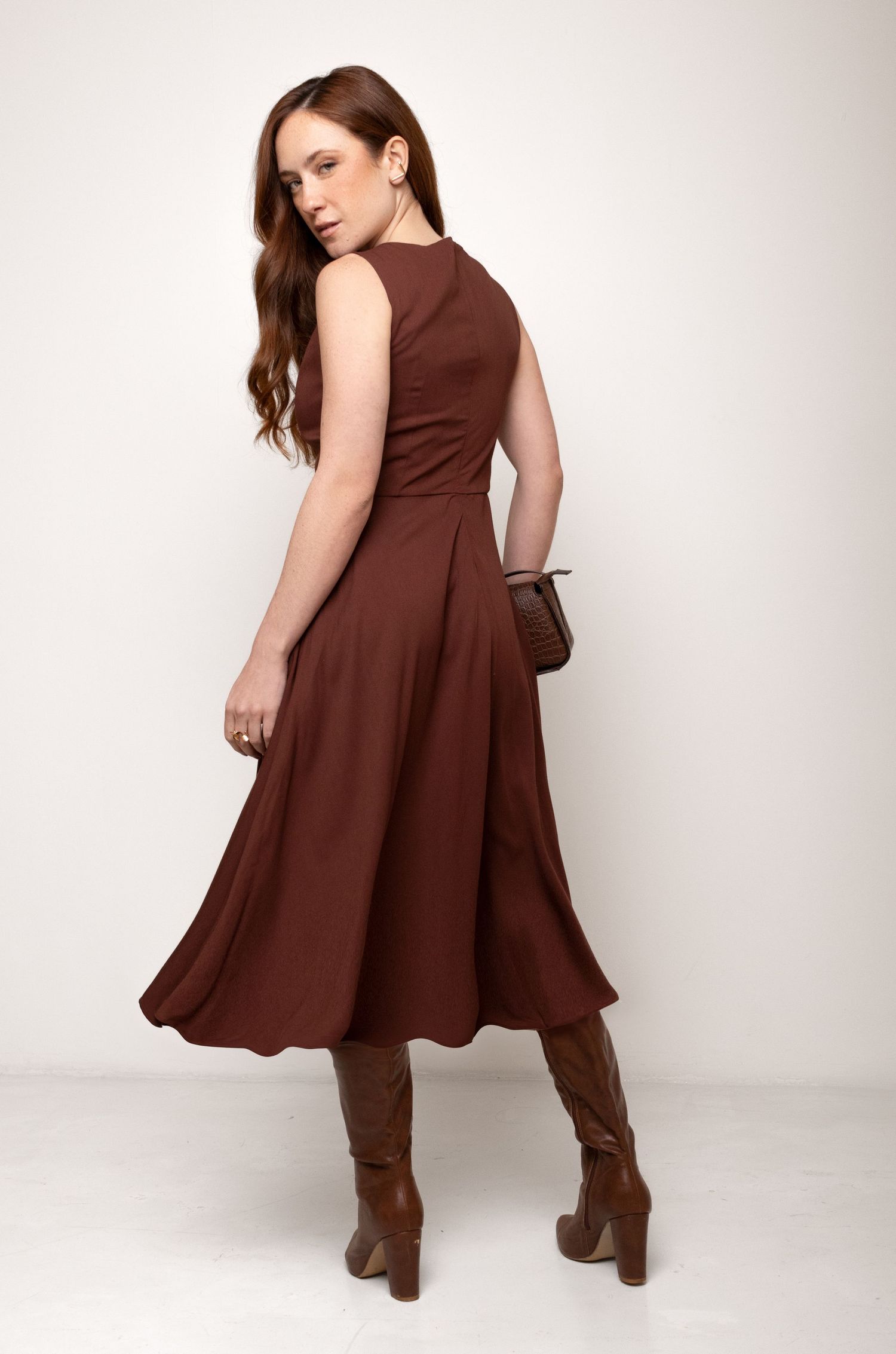 Back view of a woman in a sleeveless brown midi dress with a flowing skirt, paired with tall brown boots and a small clutch.