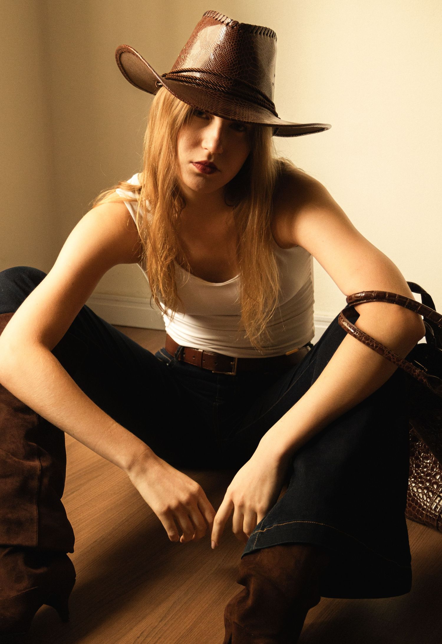 Woman with long blonde hair in a brown leather cowboy hat, white tank top, dark jeans and suede boots sits on a wooden floor in warm light.