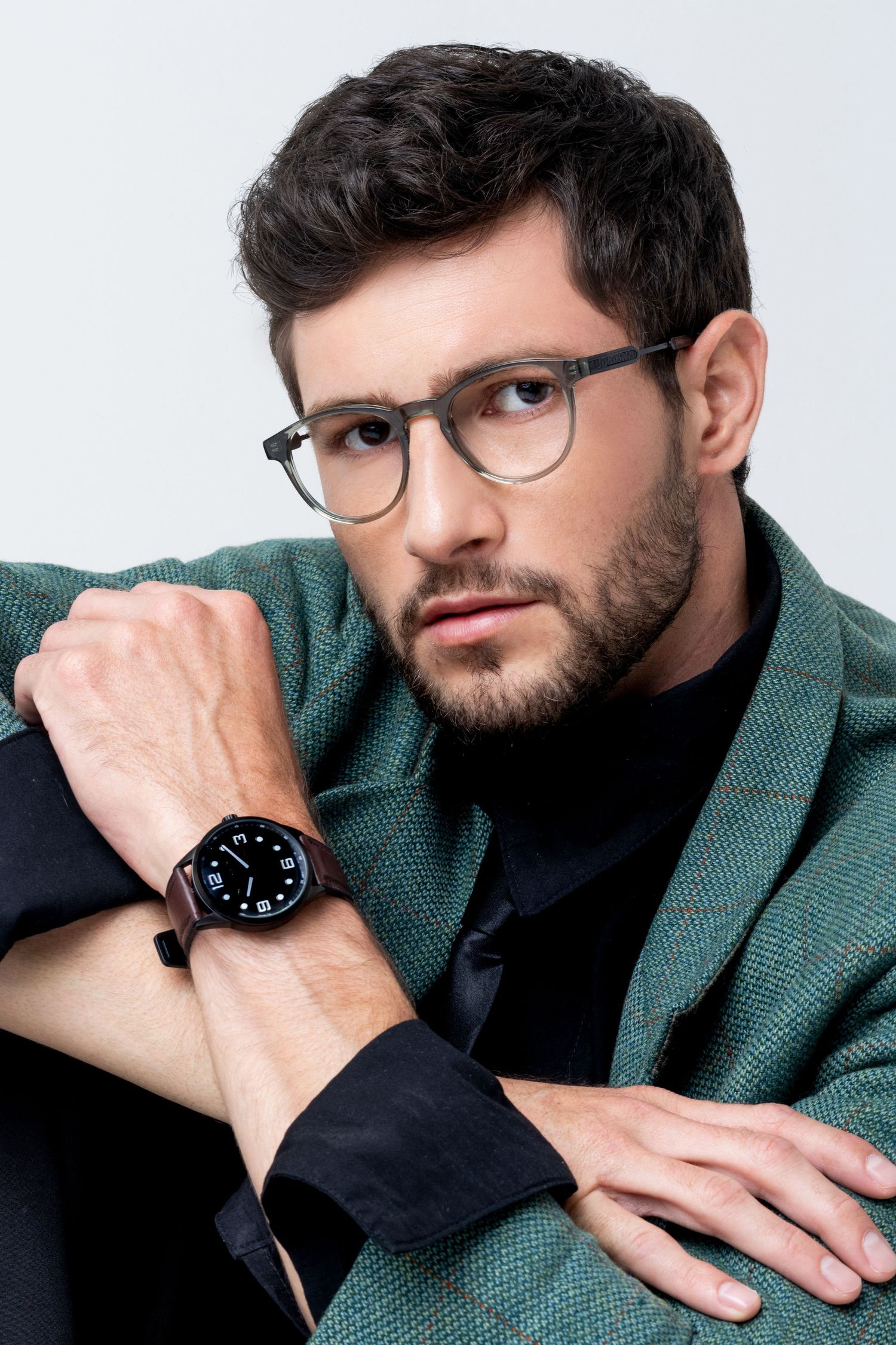 Bearded man in clear-frame glasses and a green tweed blazer rests his arm on his knee, showing a black dial watch with brown strap.