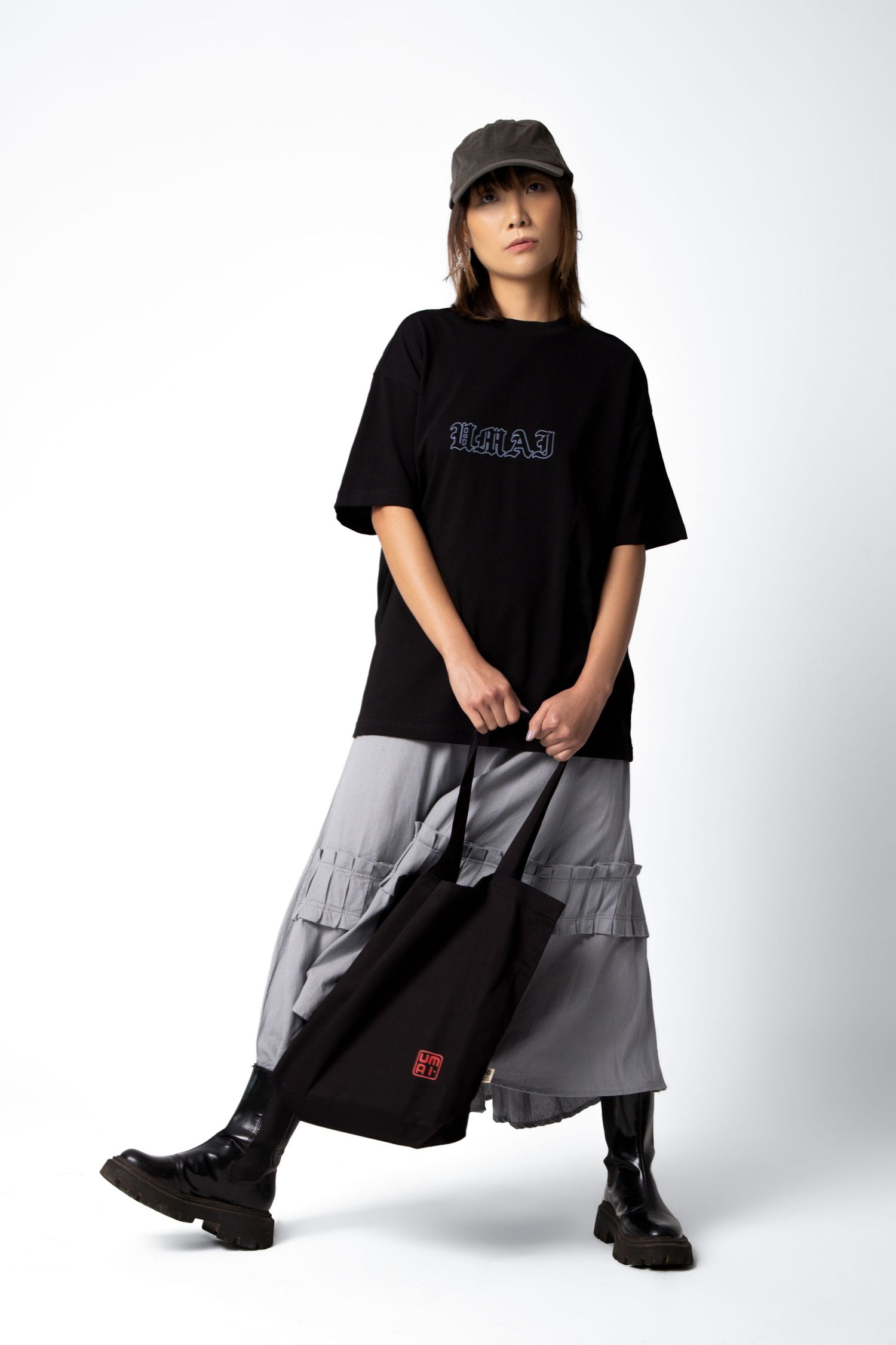 Woman in a black graphic t-shirt, gray ruffled skirt, cap and black boots stands holding a black tote bag with a red label.