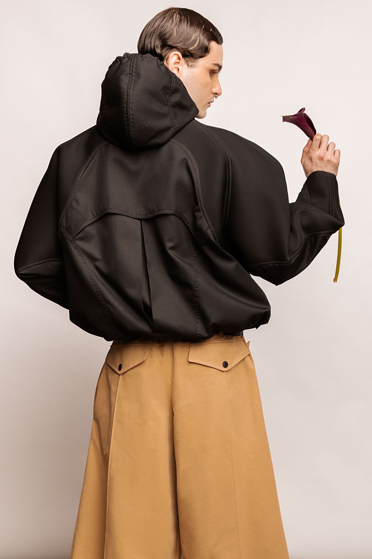 Back view of a model in an oversized black hooded jacket and tan wide-leg shorts with button flap pockets, holding a calla lily.