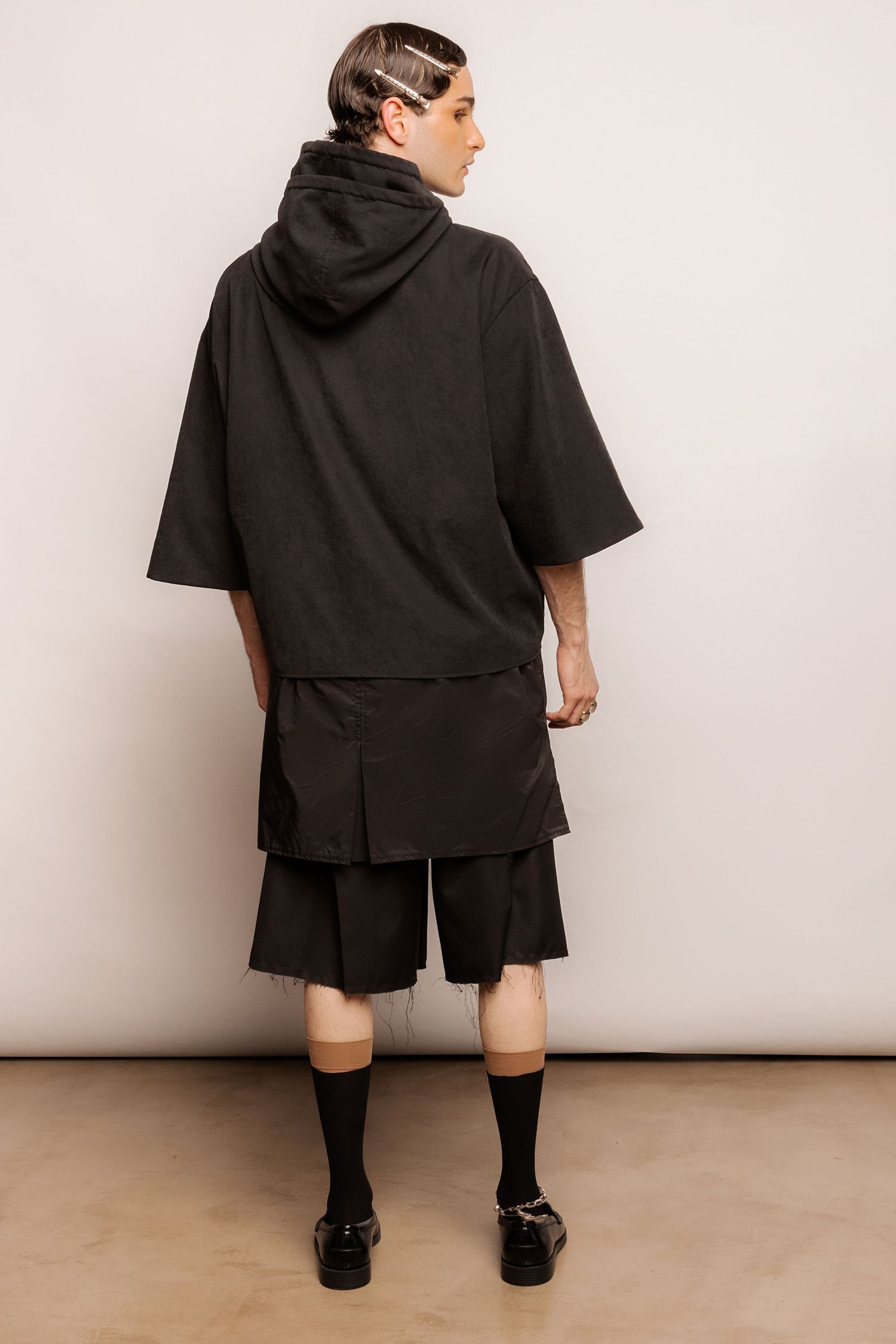 Back view of a model in an oversized black hooded top, pleated black shorts, black socks and loafers against a plain studio backdrop.