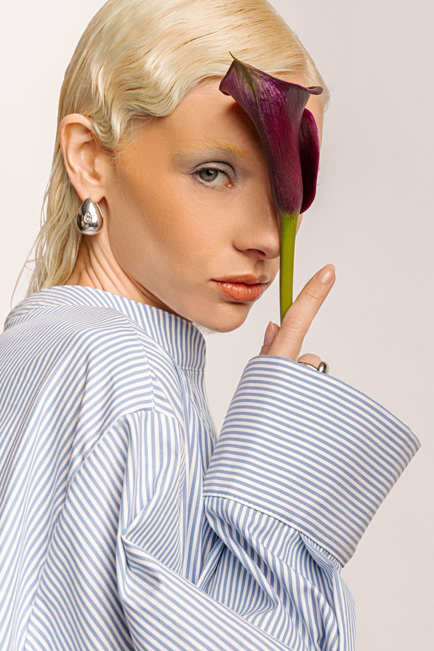Beauty close-up of a blonde model with a silver earring covering one eye with a purple calla lily, wearing a striped shirt.