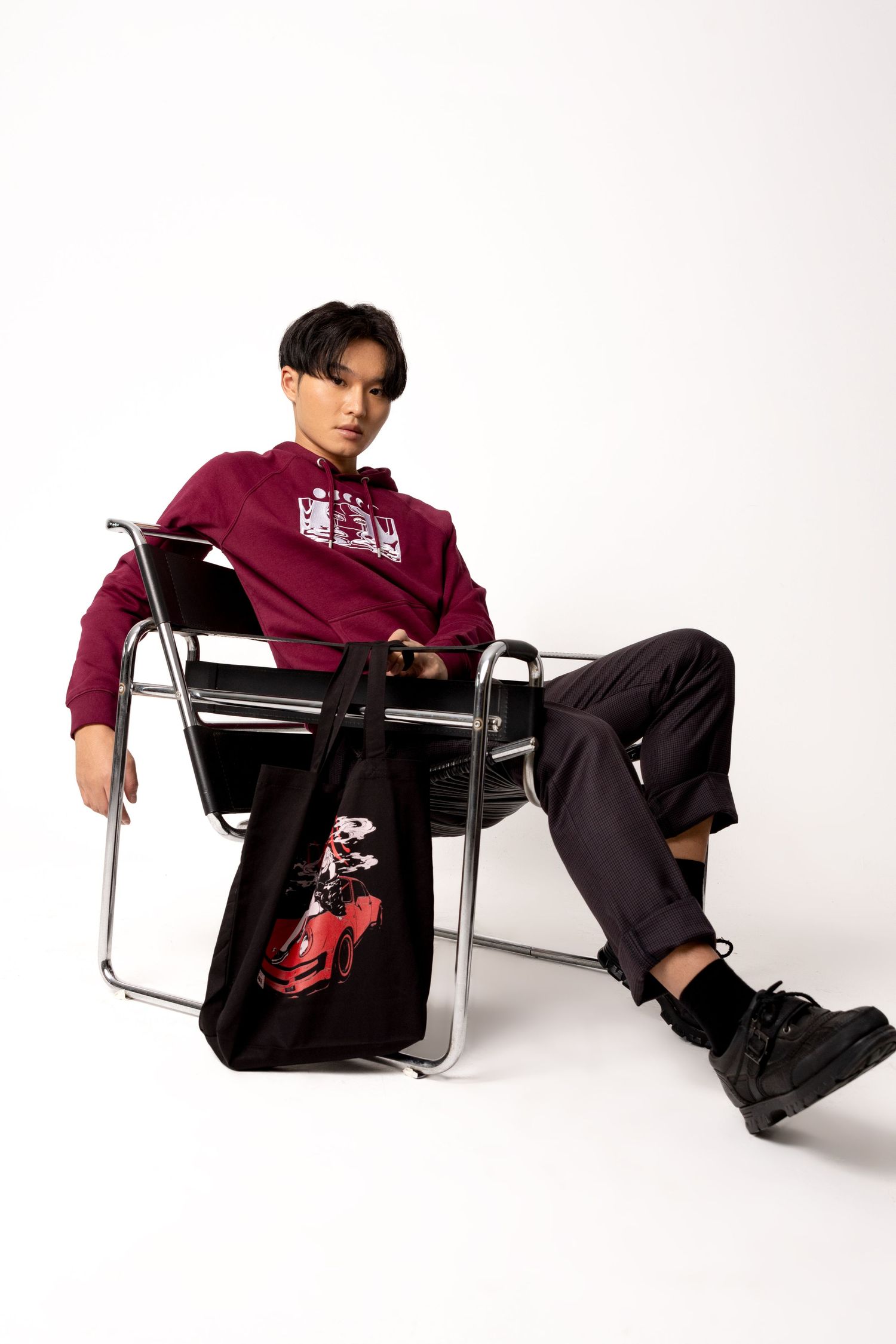 Young man in a maroon graphic hoodie and black boots sits in a chrome chair with a car print tote bag.