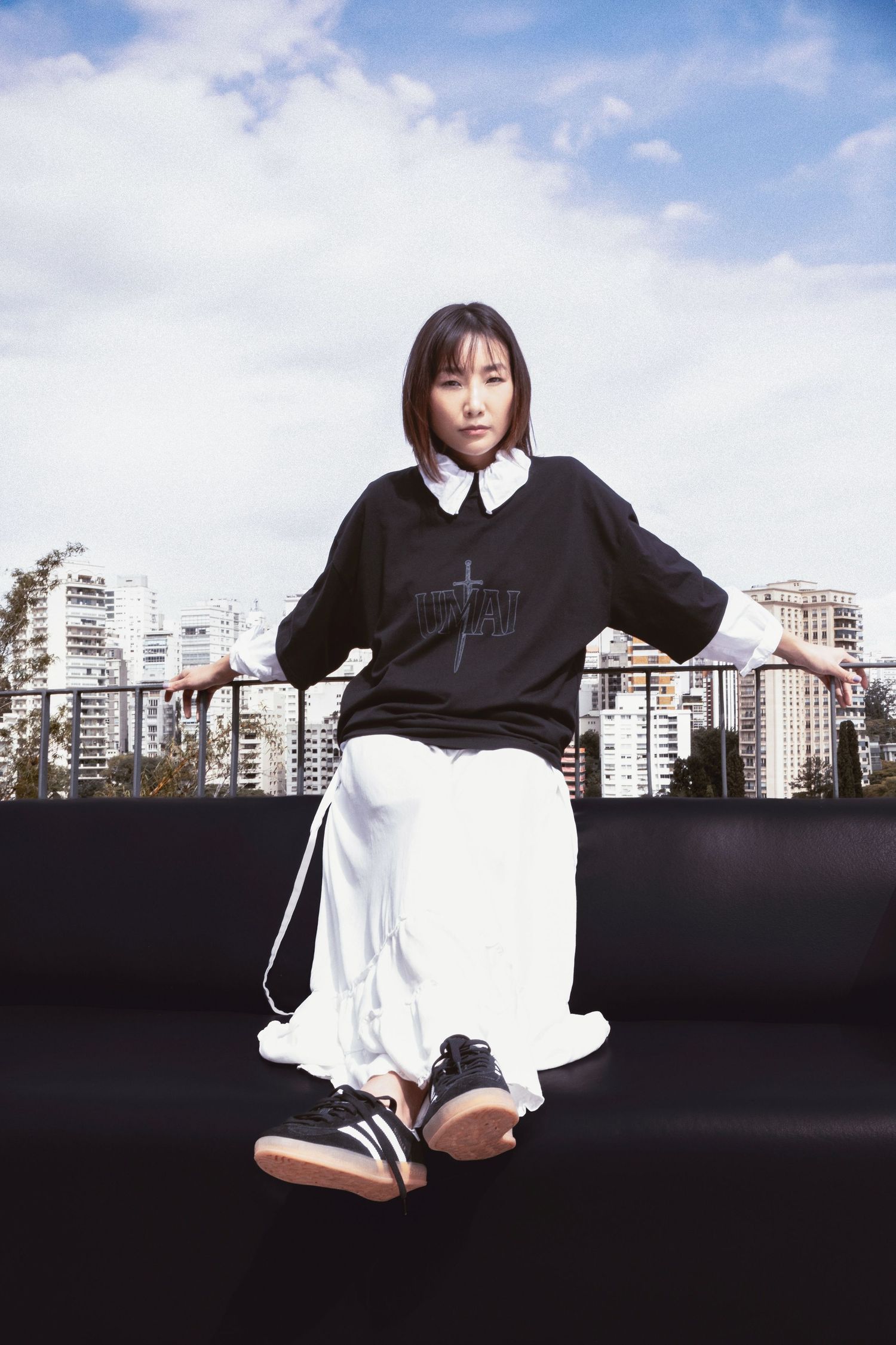 Woman in a black sweatshirt with white collar, white skirt and black sneakers sits on a black couch on a rooftop with city views.