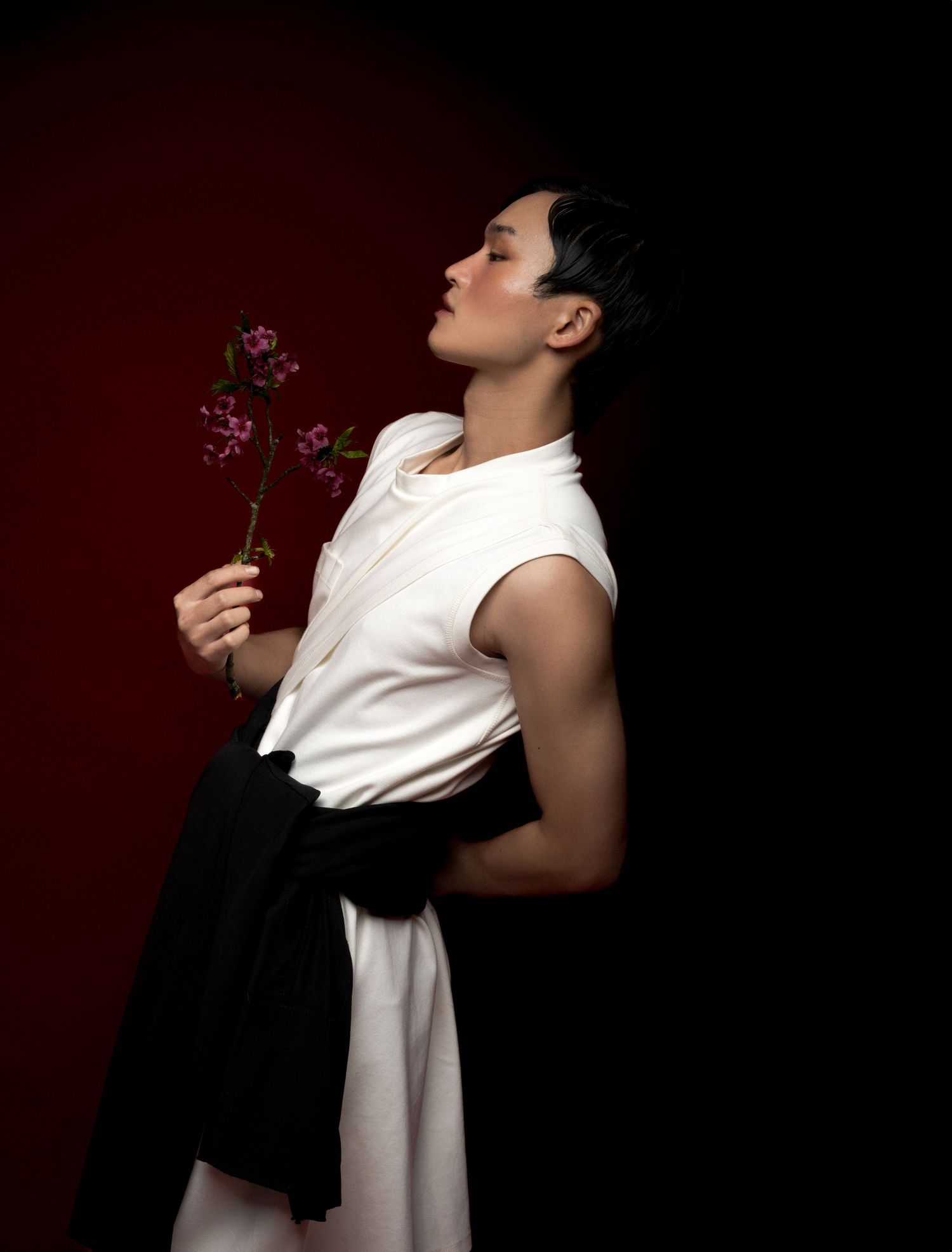 Model in a sleeveless white top with black sash holds a pink blossom branch in profile against a dramatic dark red background.