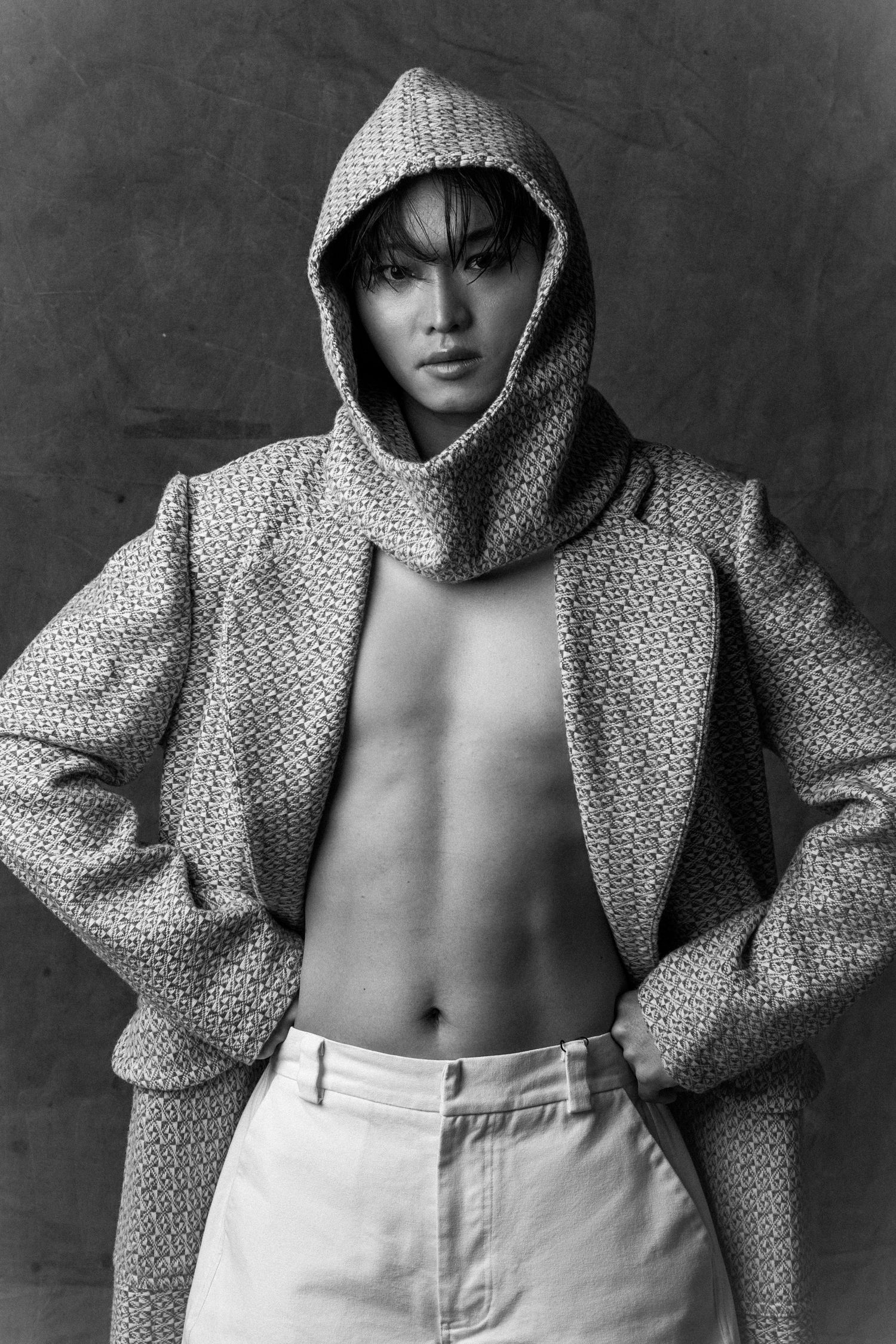 Black and white fashion portrait of a model in a hooded tweed coat and white trousers, hands on hips, with a confident gaze.