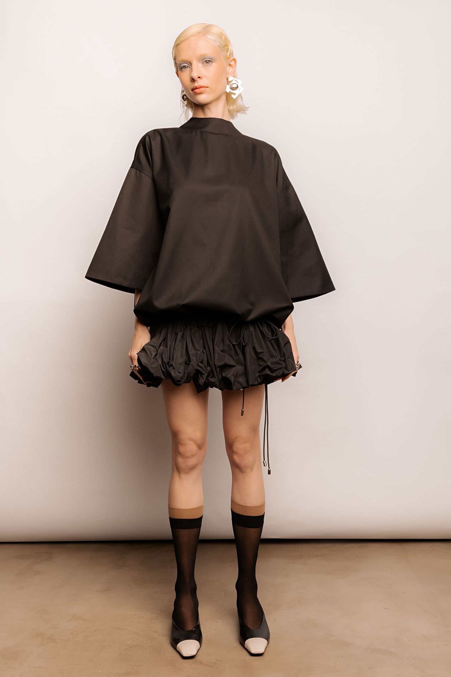 Front view of a model wearing a black oversized short-sleeve top with a gathered bubble mini skirt, sheer socks and statement earrings.