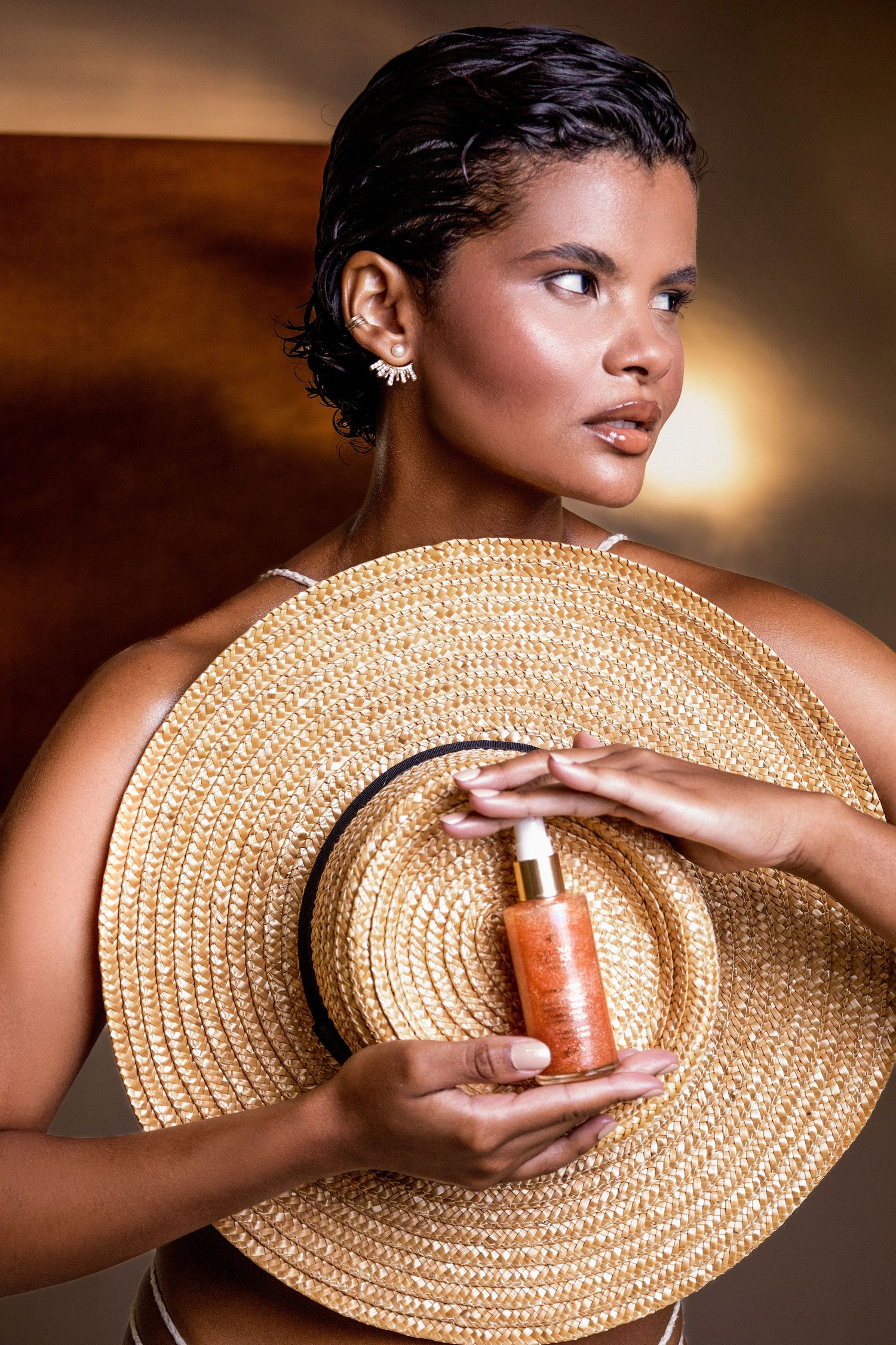 Woman with short dark hair holds a wide straw hat to her chest, displaying an orange skincare bottle with a gold dropper cap.