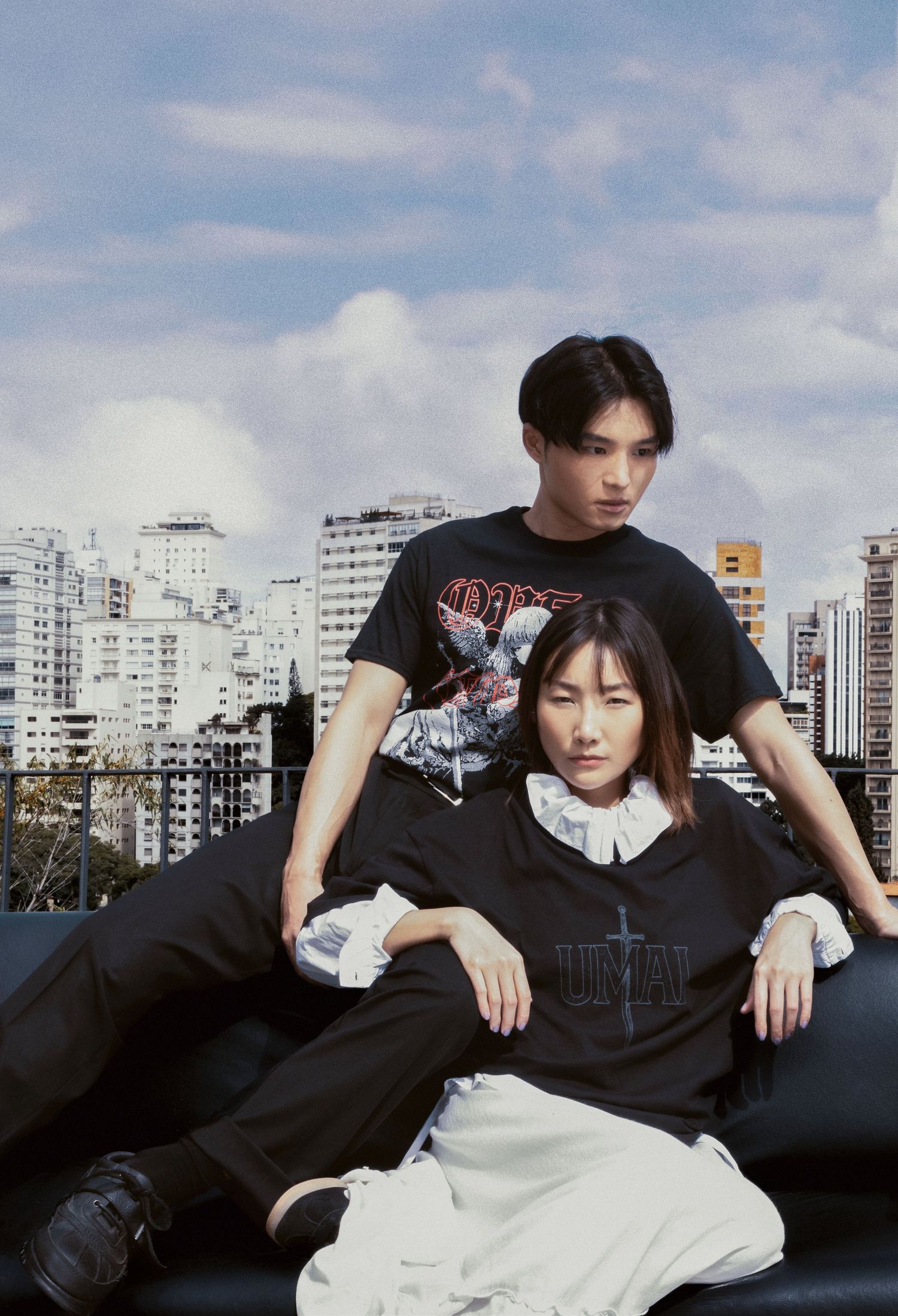 Man and woman in black streetwear with graphic tees and white collars pose together on a black couch with a city skyline behind.