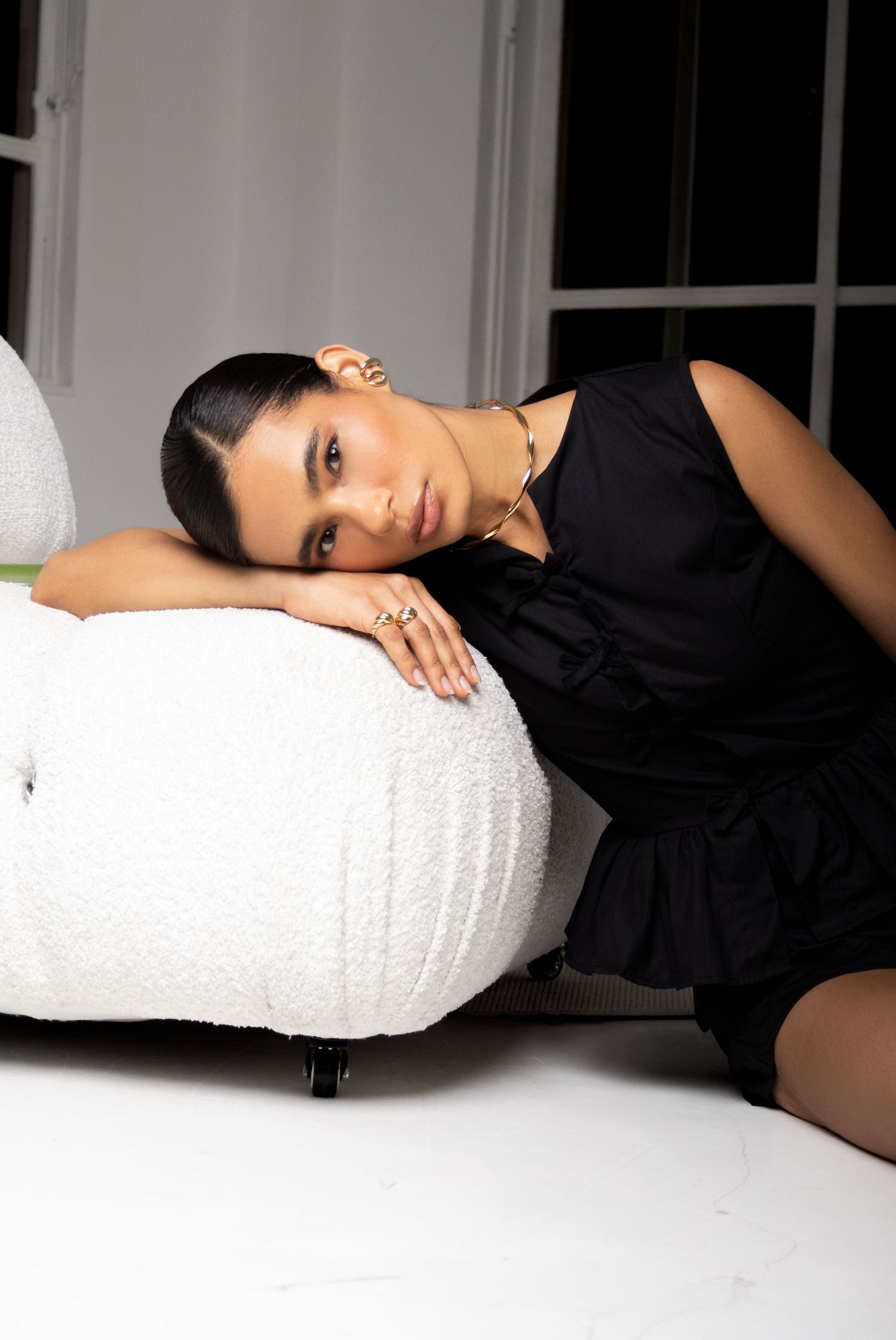 Close-up of a woman in a black sleeveless top resting her head on her arm against a white boucle sofa near large windows.