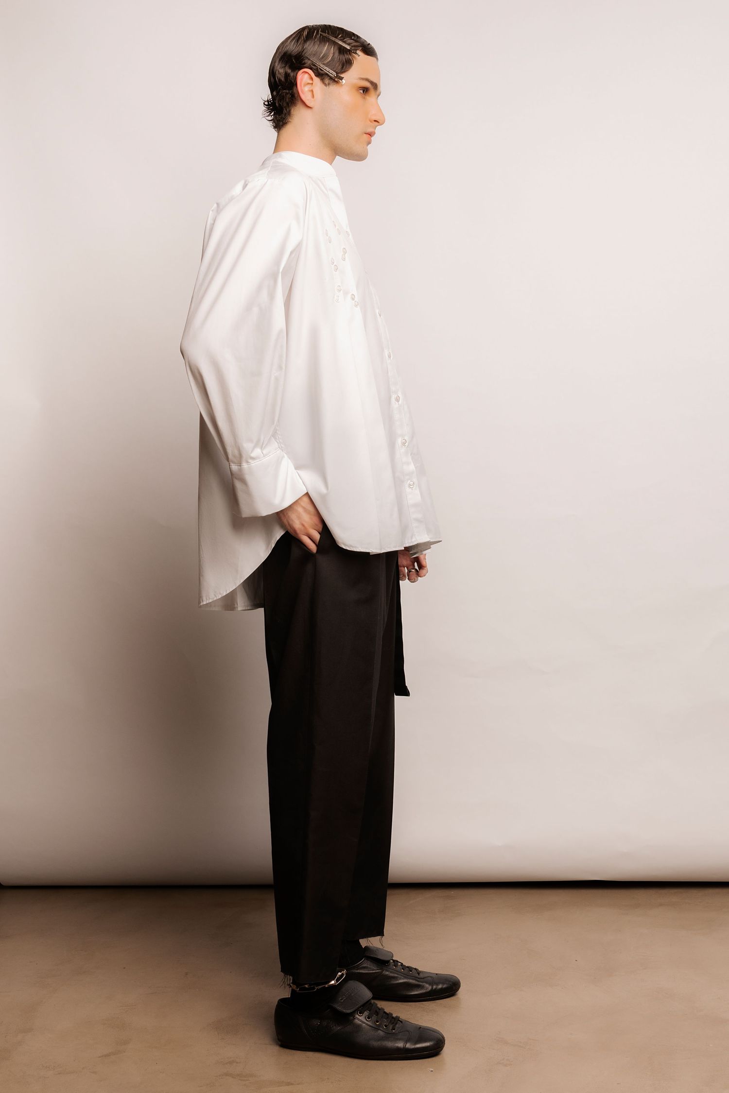 Side view of a model in a loose white collarless shirt with rolled cuffs, black belted trousers and black leather shoes, hand in pocket.