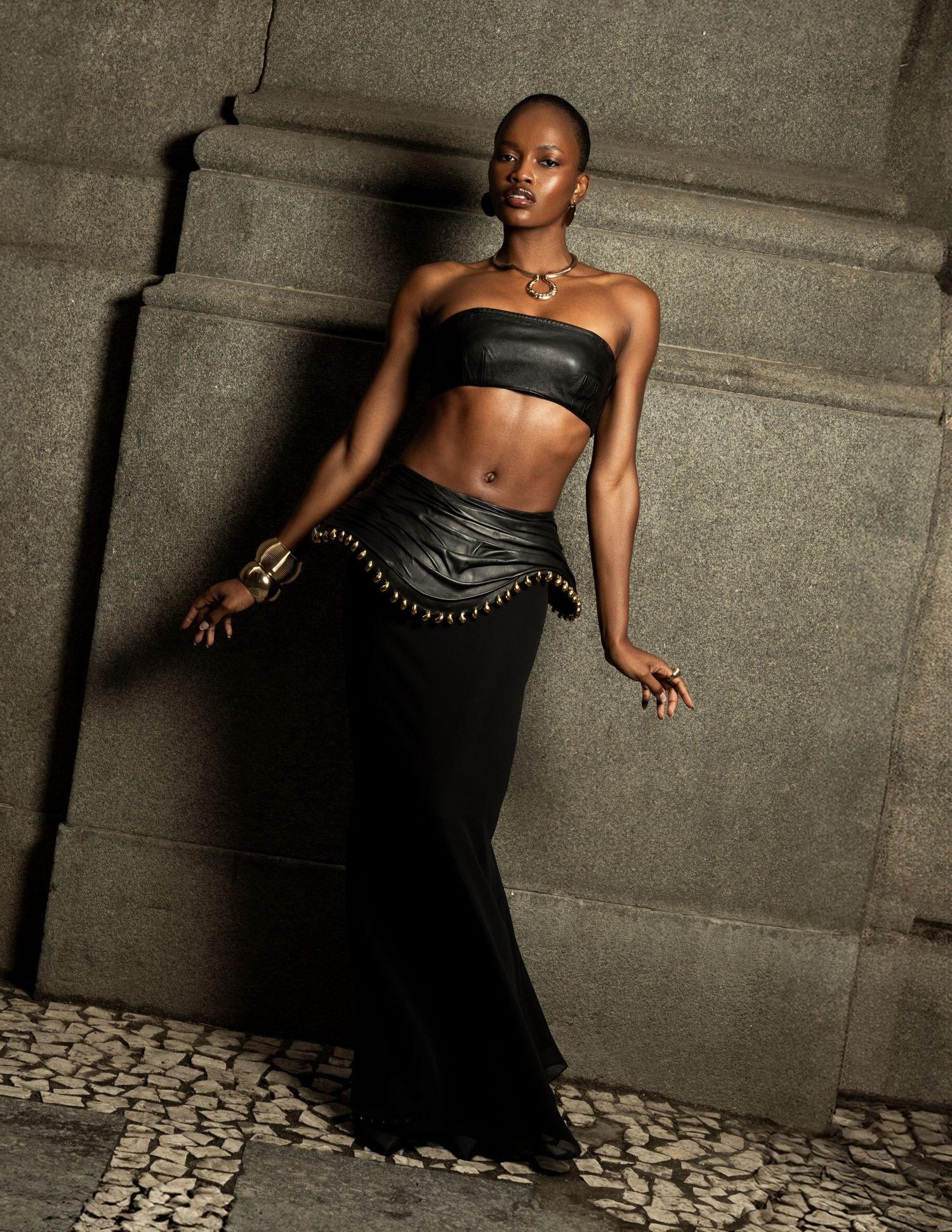 Woman in a black leather bandeau, studded belt and long black skirt leans against a stone wall on a cobbled pavement.