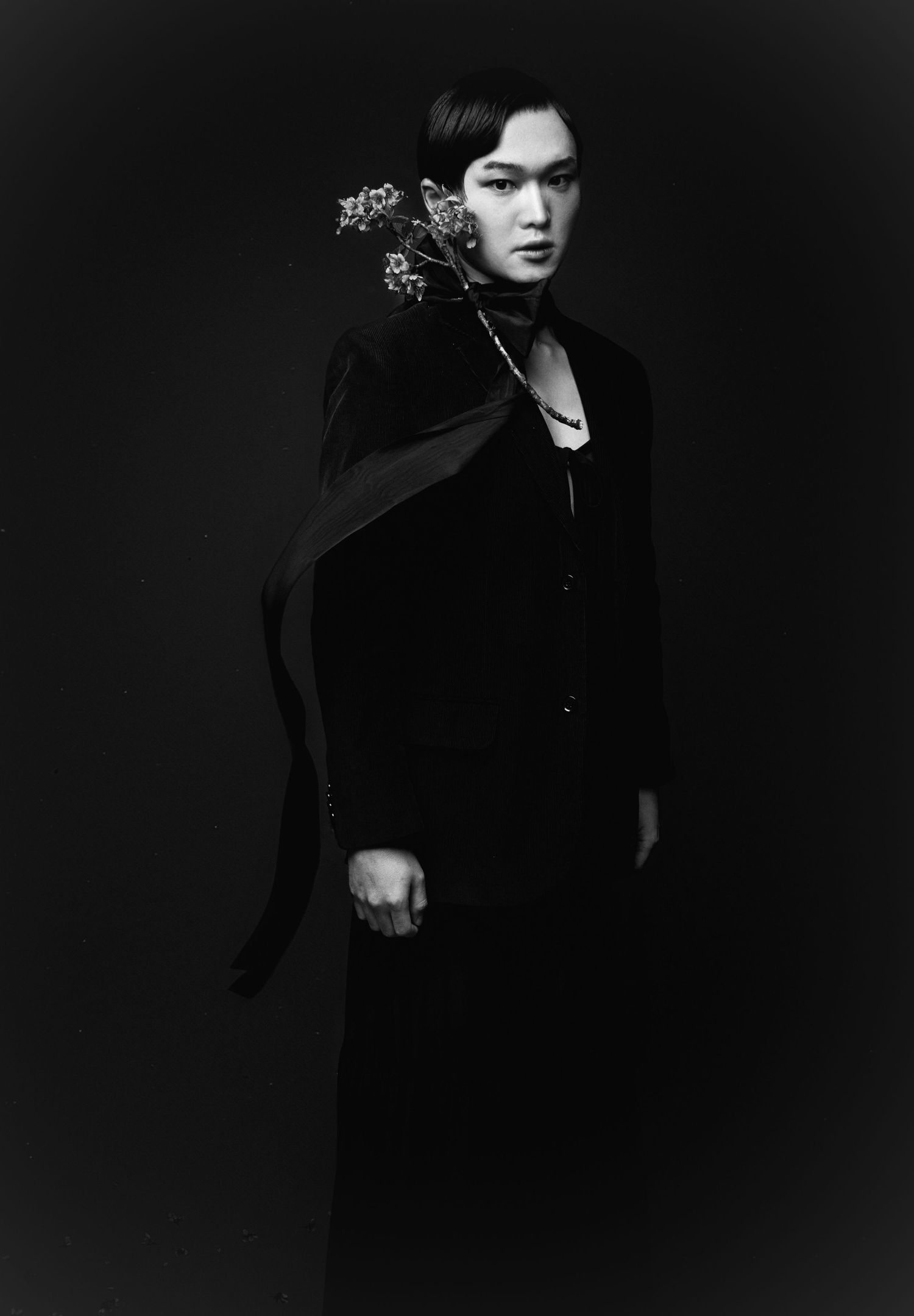Moody black and white fashion portrait of a model in dark layered clothing with a flowing scarf and flower sprigs at the shoulder.