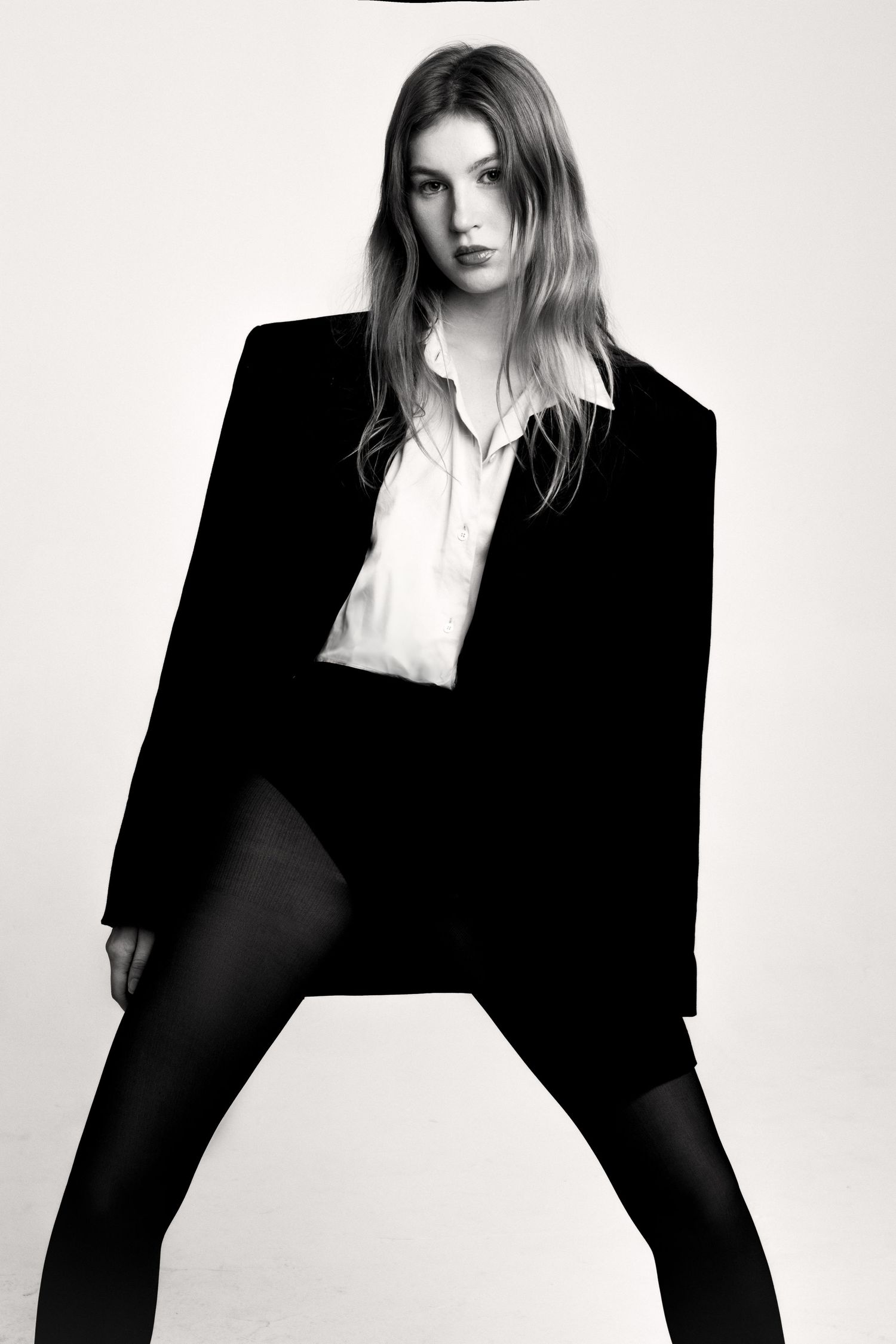 Black and white fashion portrait of a woman in an oversized black blazer, white shirt and black tights standing with legs apart.
